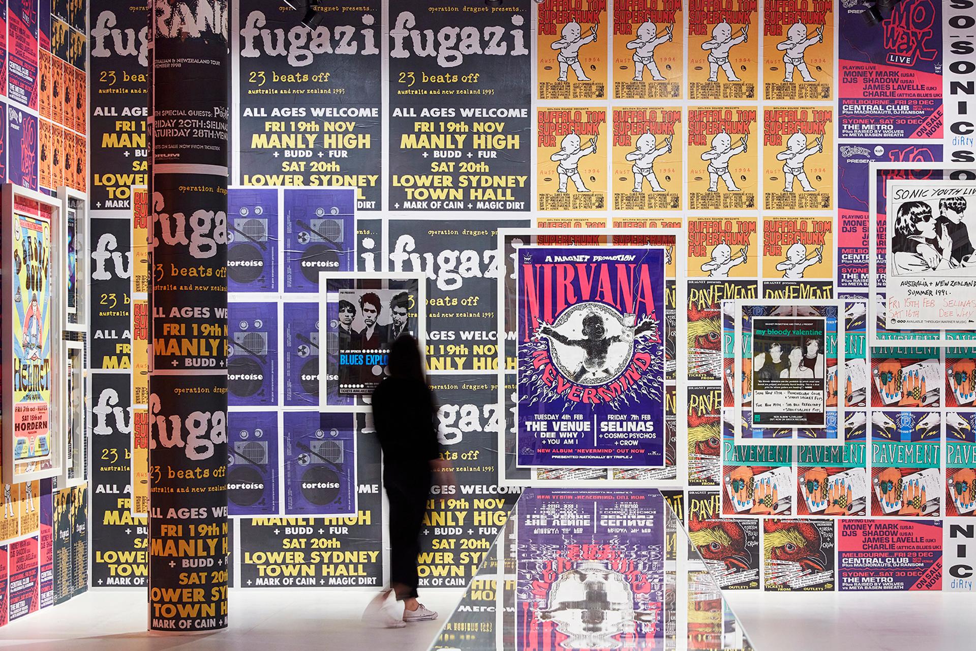 A large wall covered in a variety of brightly coloured band posters. A woman dressed in black is looking at the wall.