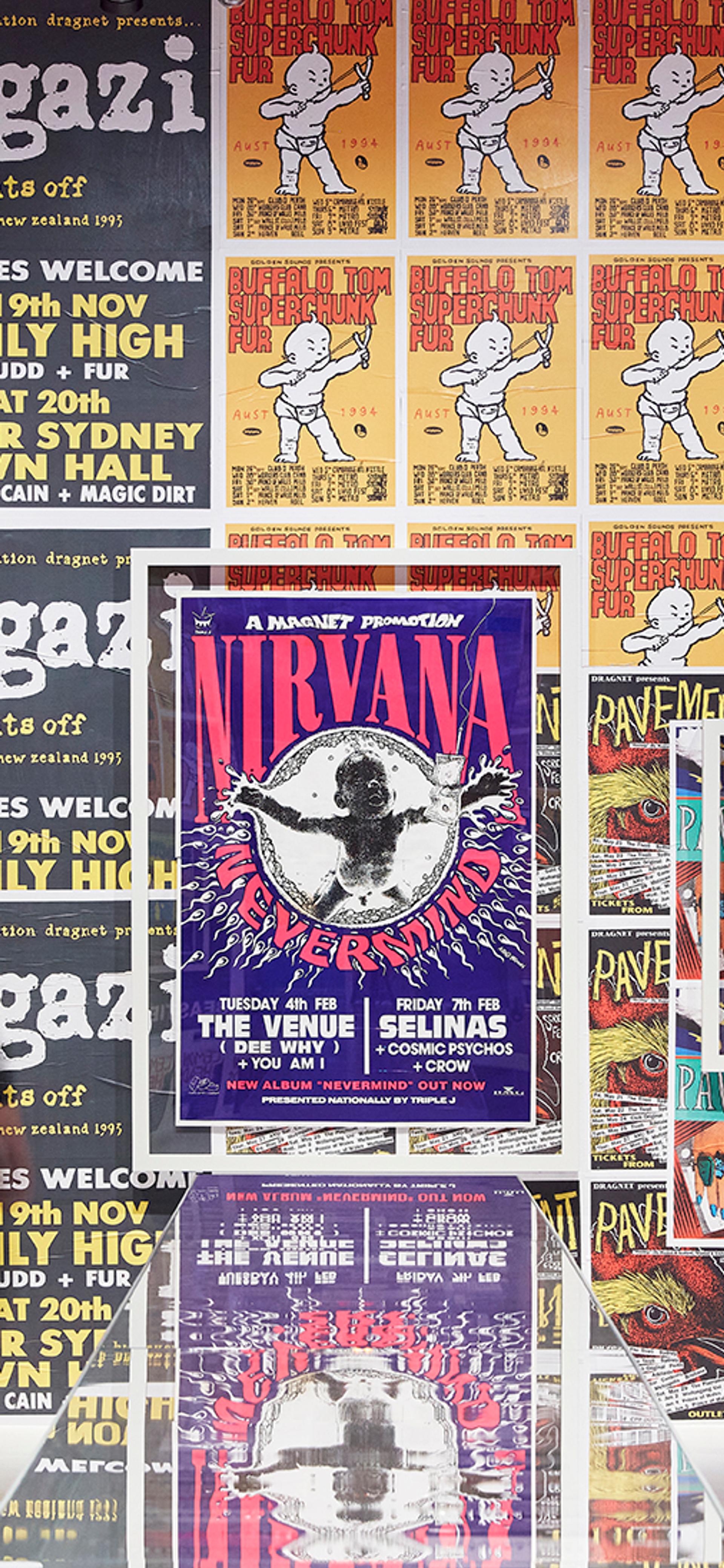 A large wall covered in a variety of brightly coloured band posters. A woman dressed in black is looking at the wall.