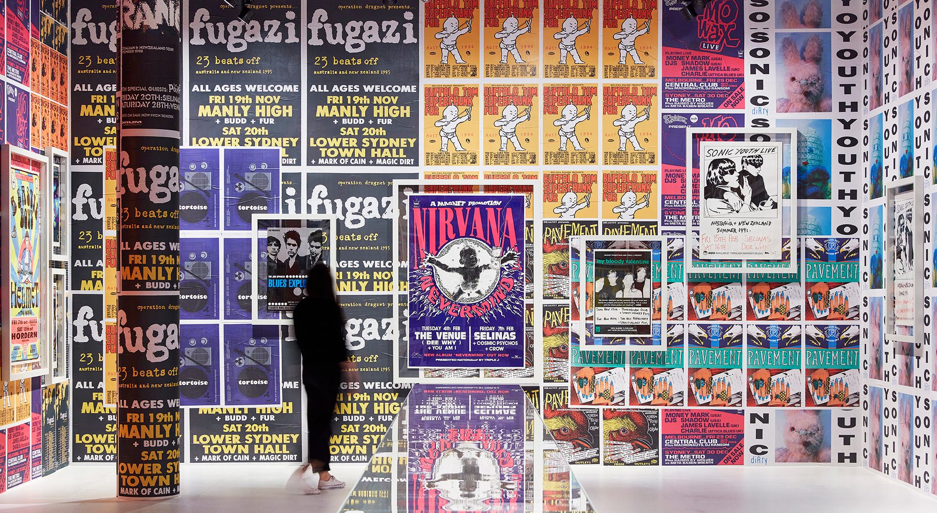 A large wall covered in a variety of brightly coloured band posters. A woman dressed in black is looking at the wall.
