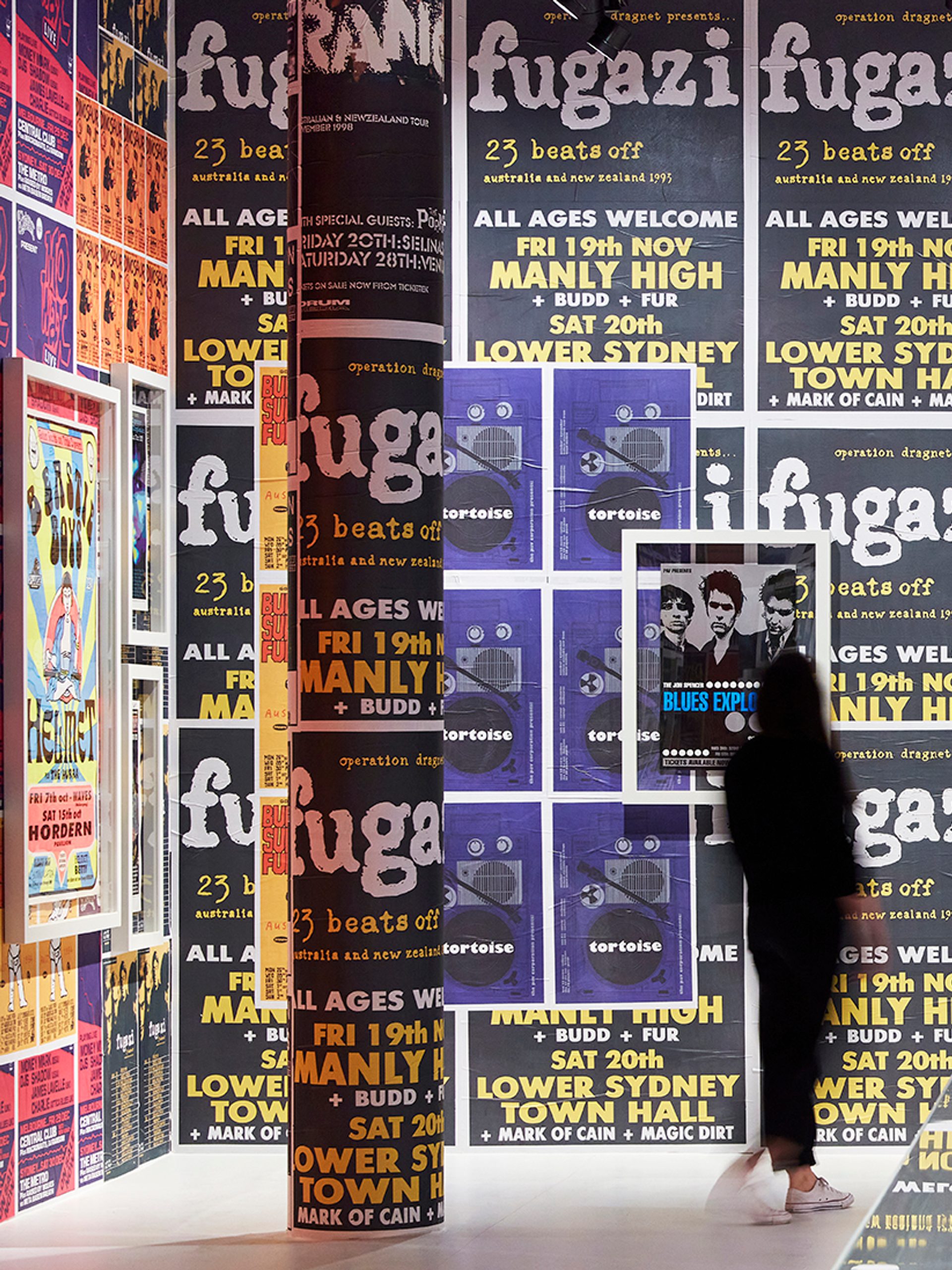 A large wall covered in a variety of brightly coloured band posters. A woman dressed in black is looking at the wall.
