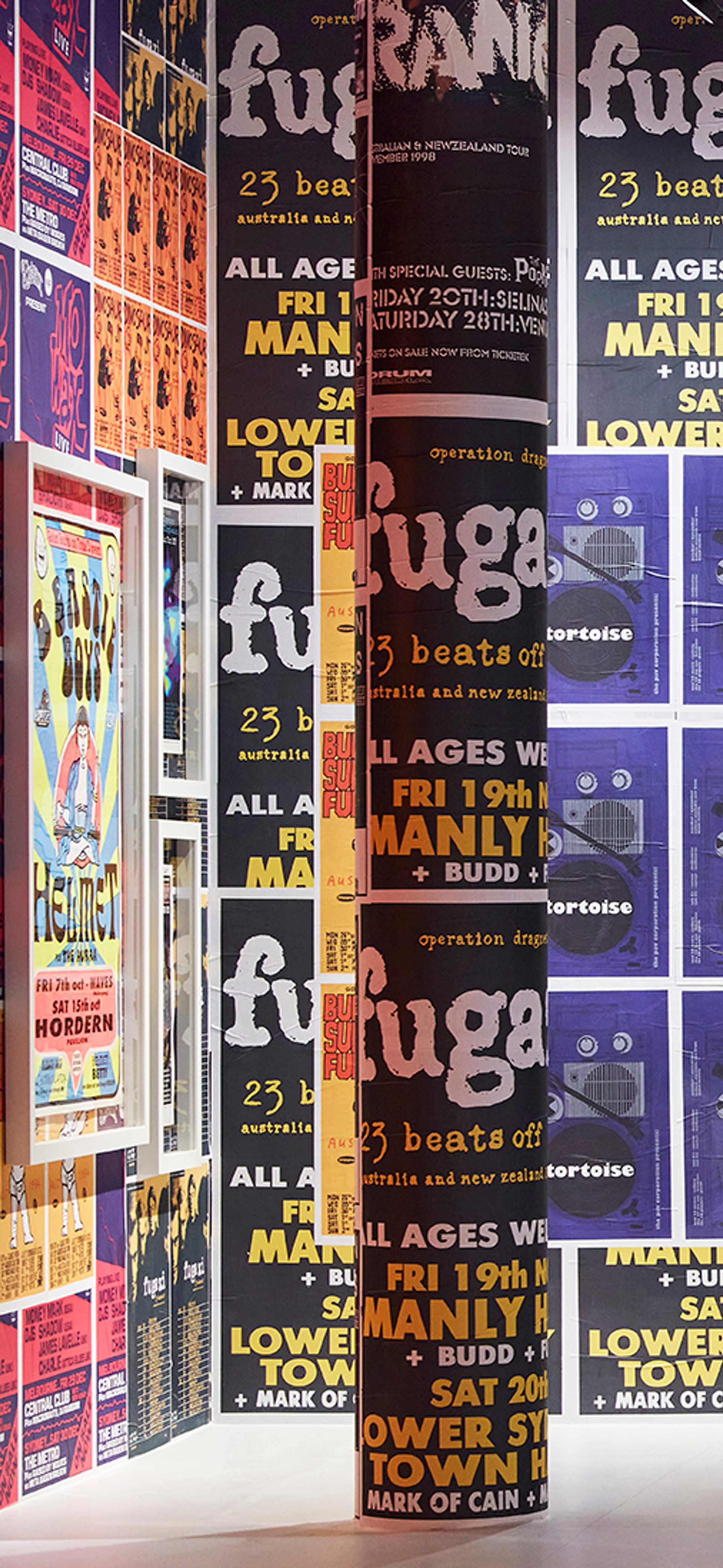 A large wall covered in a variety of brightly coloured band posters. A woman dressed in black is looking at the wall.