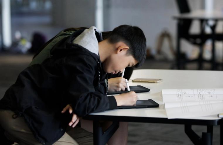 A Creative Studio participant wearing a black hoodie leans over a table and is drawing.