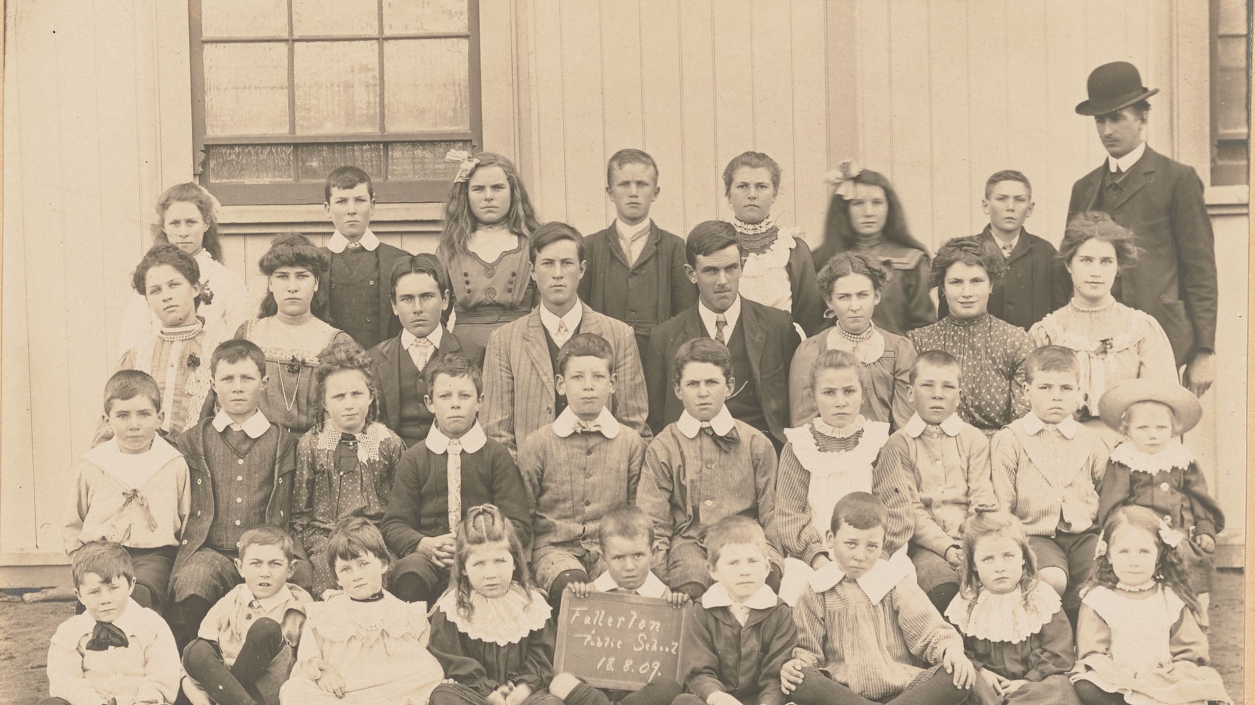 Photograph of a group study of "Fullerton Public School" pupils, posed in four rows in front of a wooden building with man in suit and bowler hat. There are 34 children of ages ranging from about 5 to about 15. The boy at centre front is holding a sign reading 'Fullerton / Public School / 18.8.09.'. Horizontal format on embossed cream mount.
