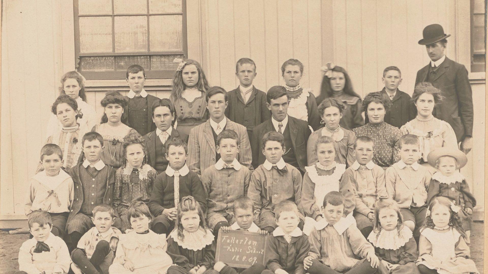 Photograph of a group study of "Fullerton Public School" pupils, posed in four rows in front of a wooden building with man in suit and bowler hat. There are 34 children of ages ranging from about 5 to about 15. The boy at centre front is holding a sign reading 'Fullerton / Public School / 18.8.09.'. Horizontal format on embossed cream mount.