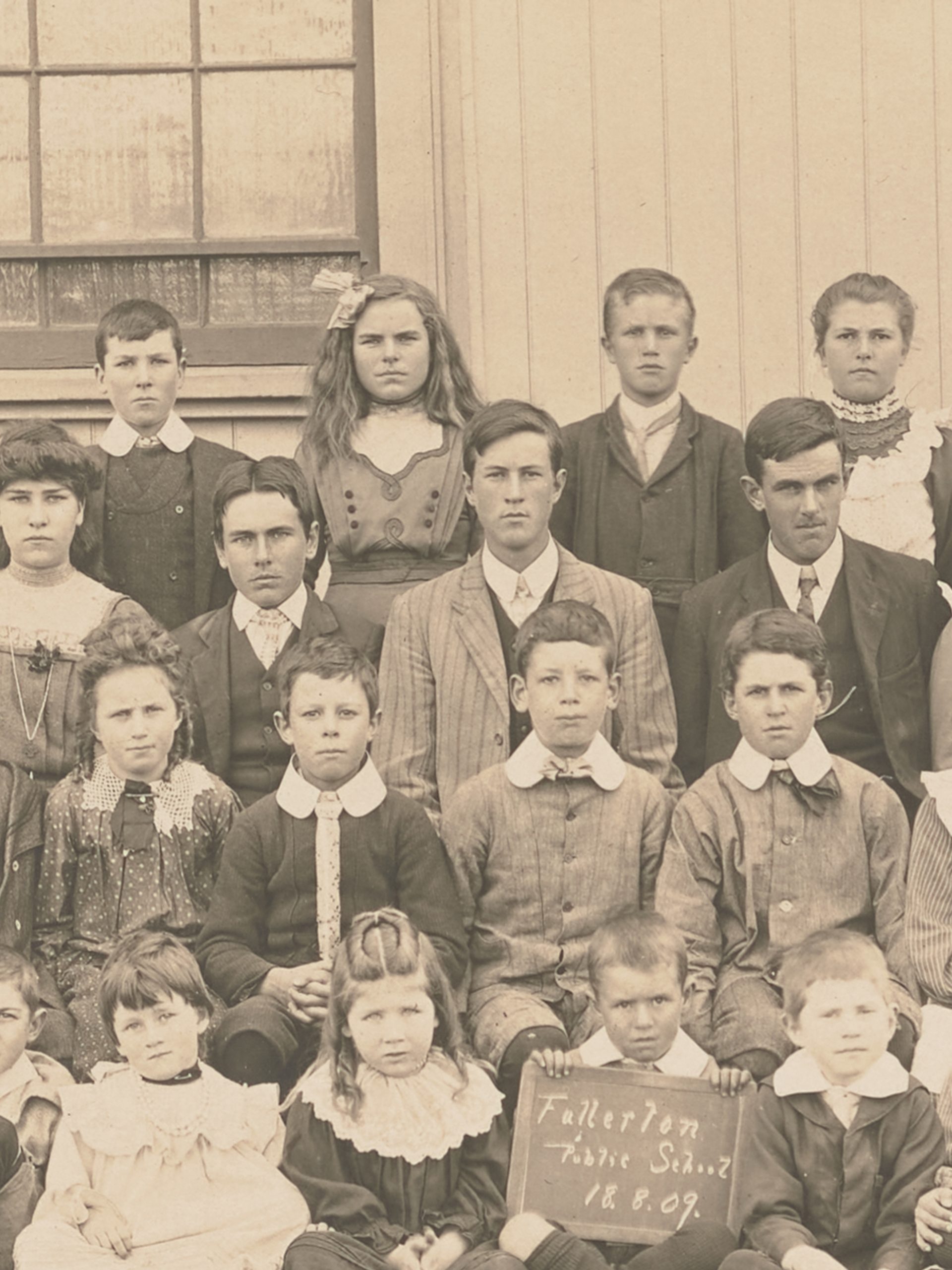 Photograph of a group study of "Fullerton Public School" pupils, posed in four rows in front of a wooden building with man in suit and bowler hat. There are 34 children of ages ranging from about 5 to about 15. The boy at centre front is holding a sign reading 'Fullerton / Public School / 18.8.09.'. Horizontal format on embossed cream mount.