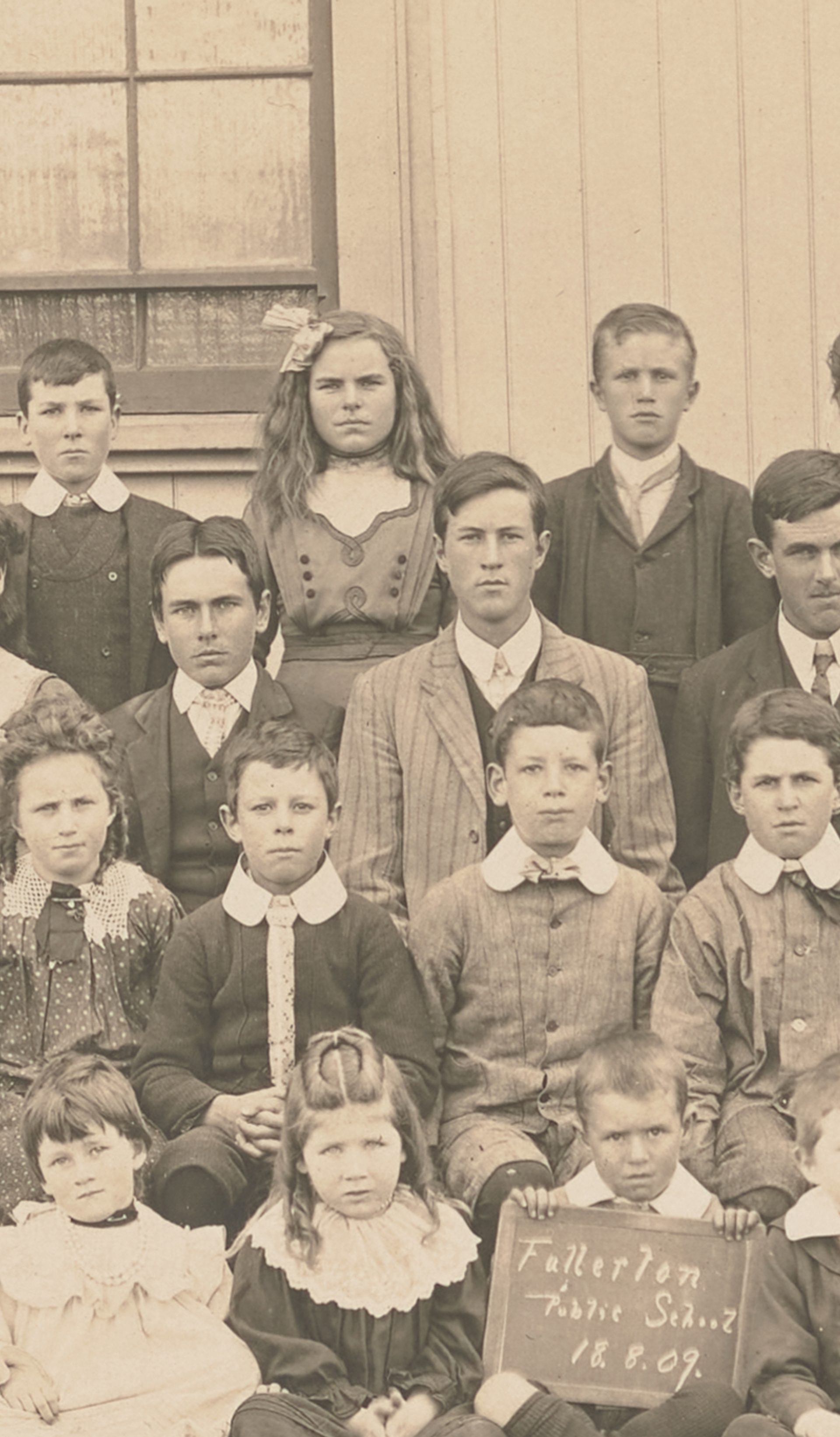 Photograph of a group study of "Fullerton Public School" pupils, posed in four rows in front of a wooden building with man in suit and bowler hat. There are 34 children of ages ranging from about 5 to about 15. The boy at centre front is holding a sign reading 'Fullerton / Public School / 18.8.09.'. Horizontal format on embossed cream mount.