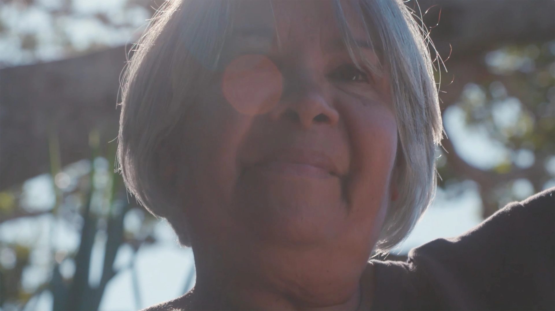 A closeup of a woman with white hair in a bob. Her dark eyes look out beyond the camera. Her right eye is obscured by a sunspot.