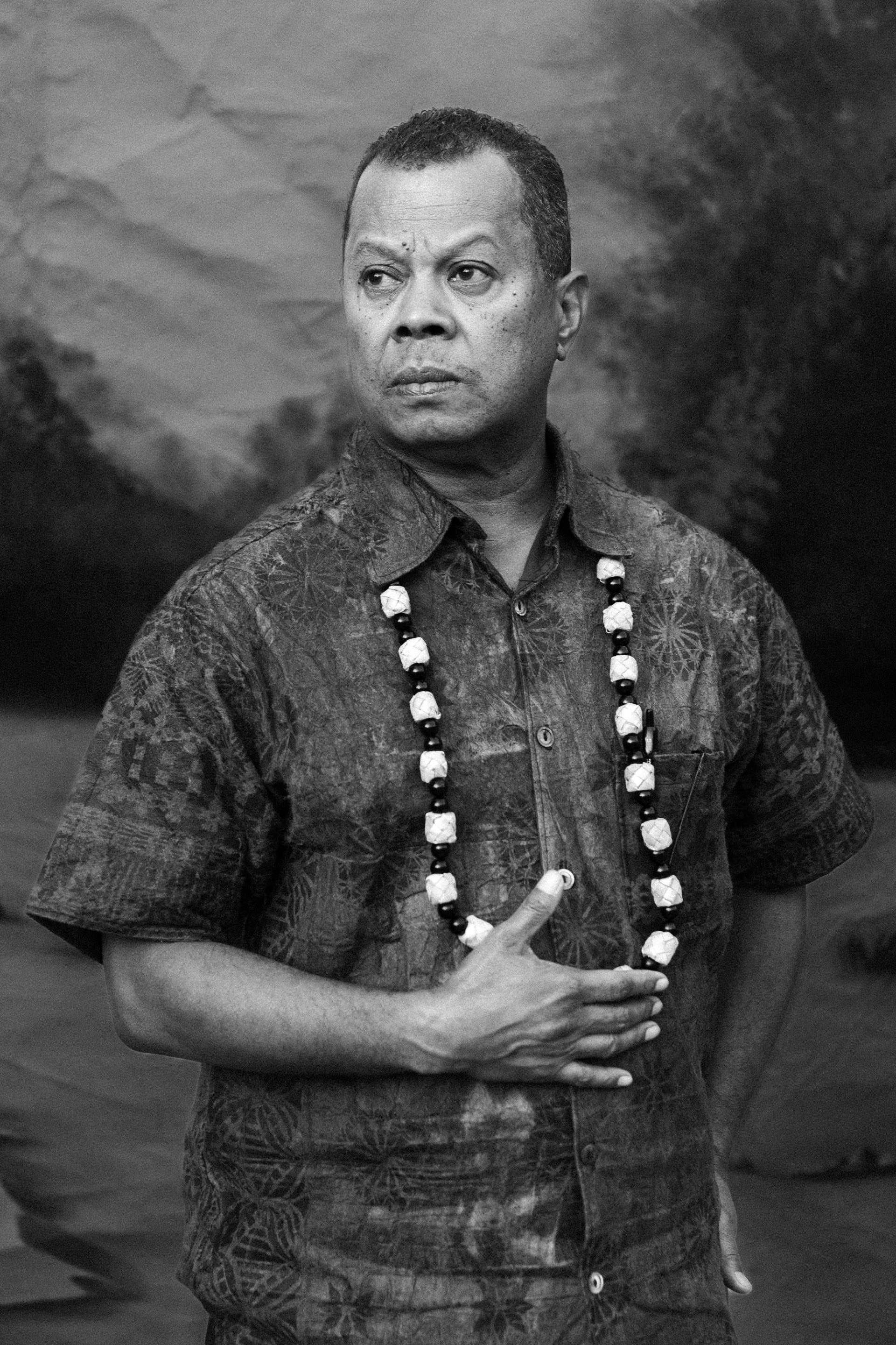 A Fijian man stands in front of a painted backdrop. He wears a shirt with a Fijian tapa print and a necklace with large beads.