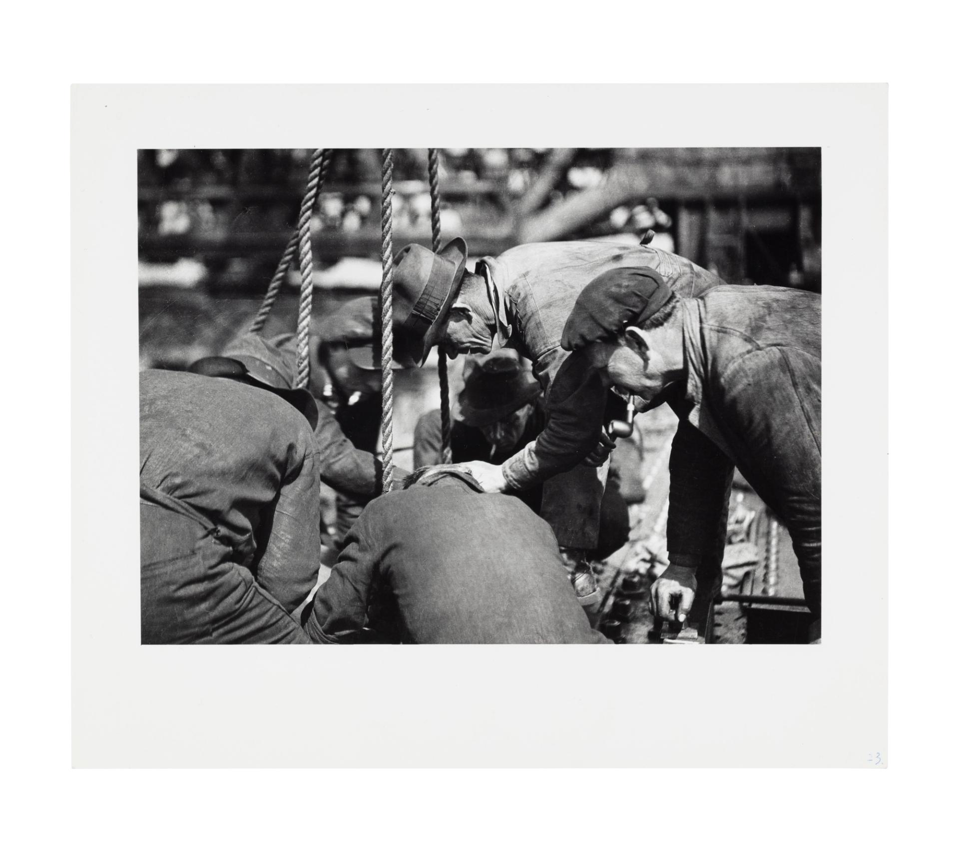 Image of six men crouching around an item or material out of frame, likely being supported or lifted by the ropes in frame. Some of the men wear hats and are smoking a pipe, they are arranged in a circle.