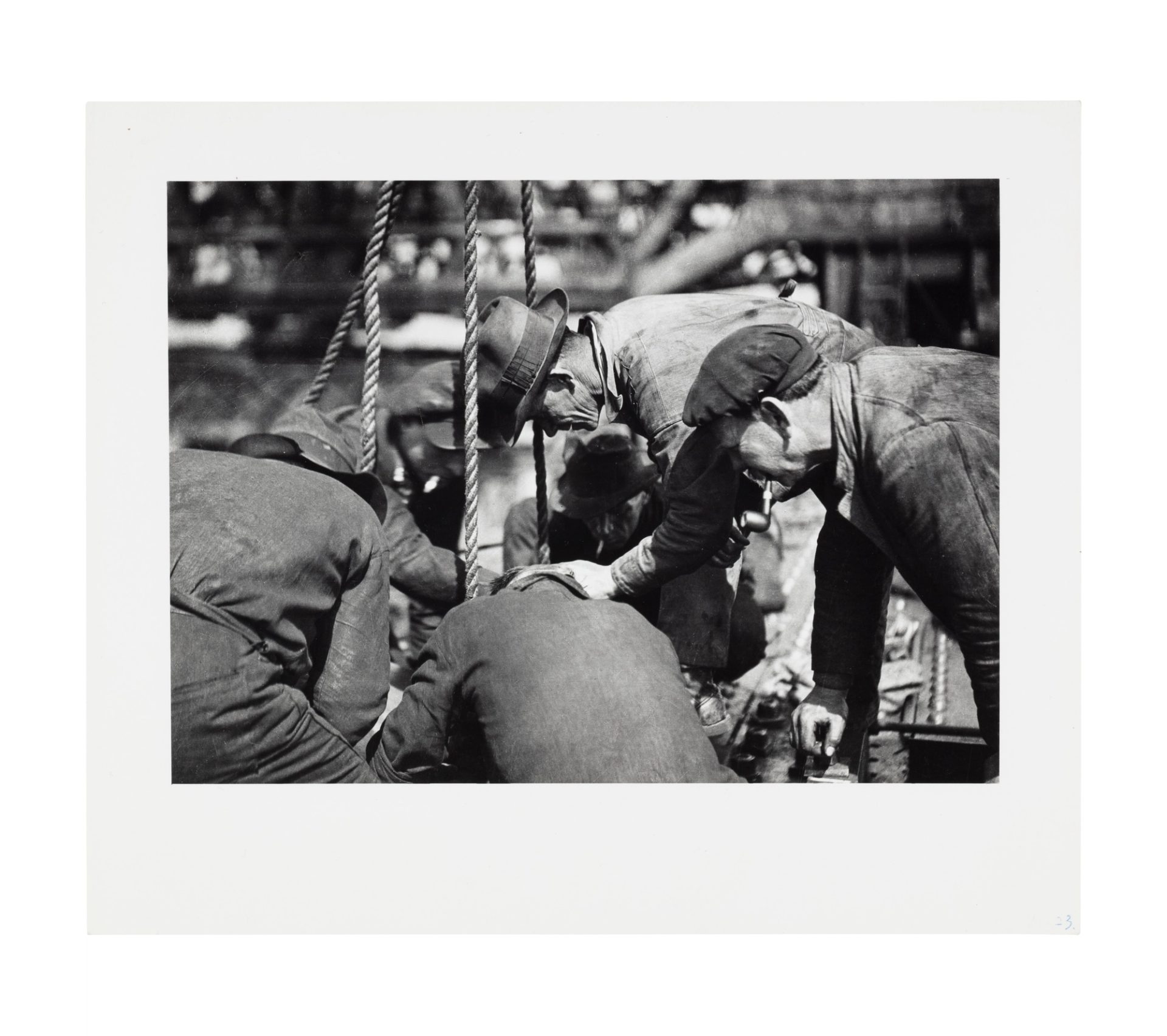 Image of six men crouching around an item or material out of frame, likely being supported or lifted by the ropes in frame. Some of the men wear hats and are smoking a pipe, they are arranged in a circle.