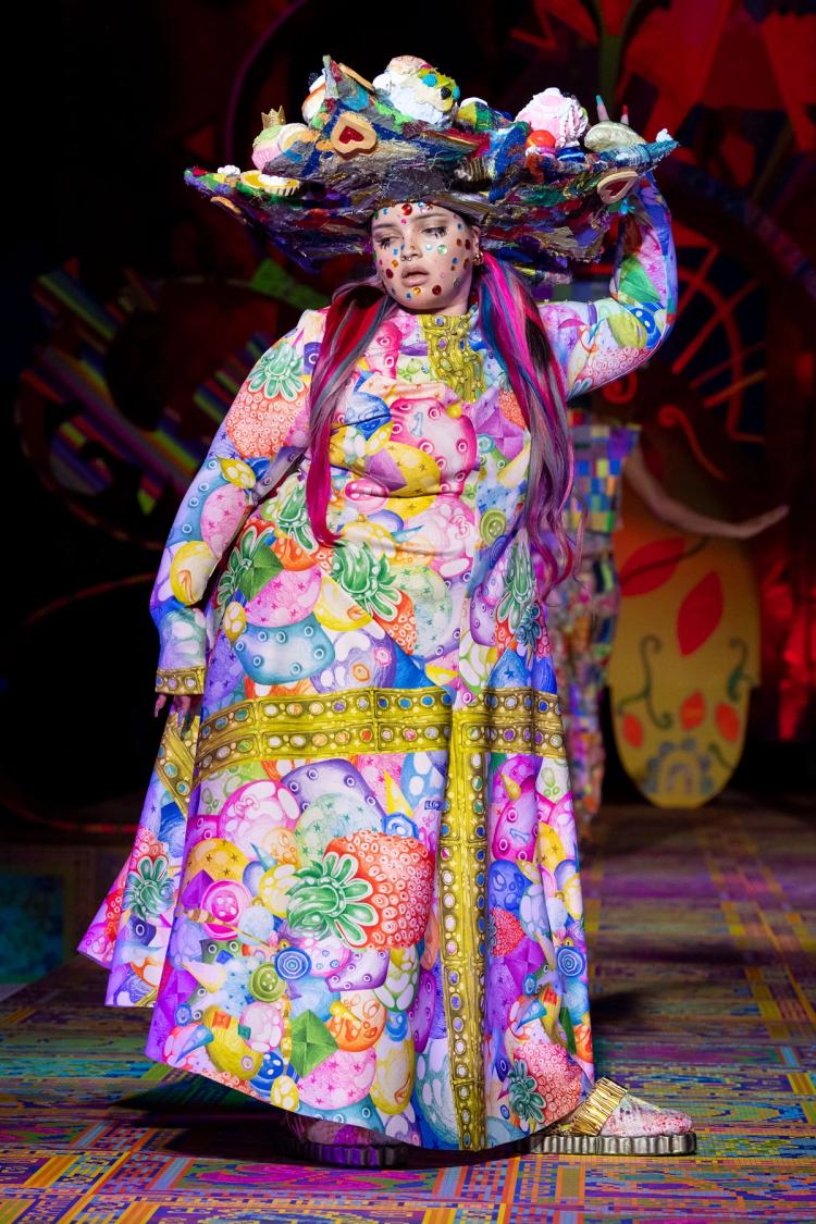 Runway model wearing colourful full length dress with a large hat and gemstone stickers covering their face.