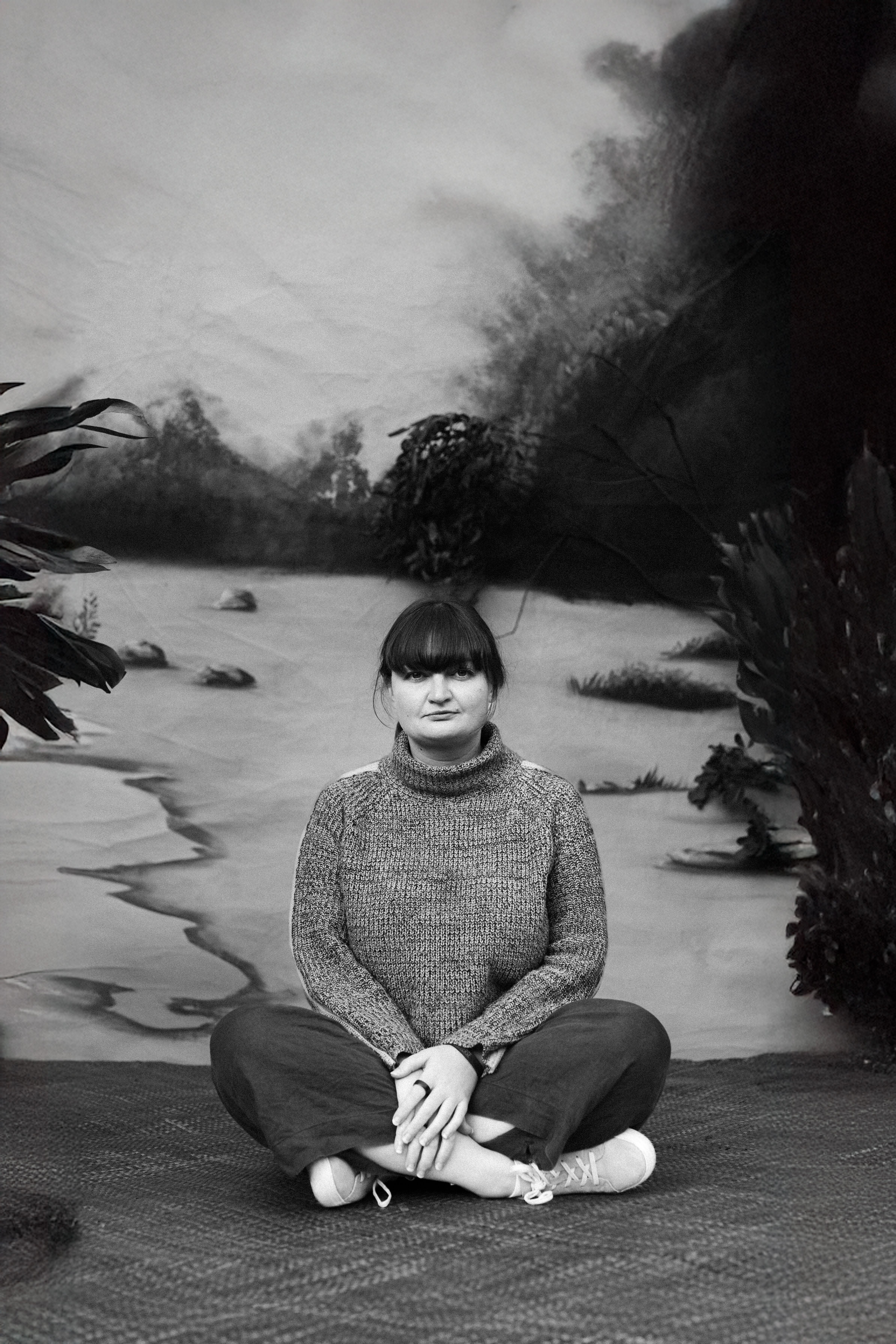 A Palawa woman sits cross-legged on a woven mat. There is a painted backdrop behind her.