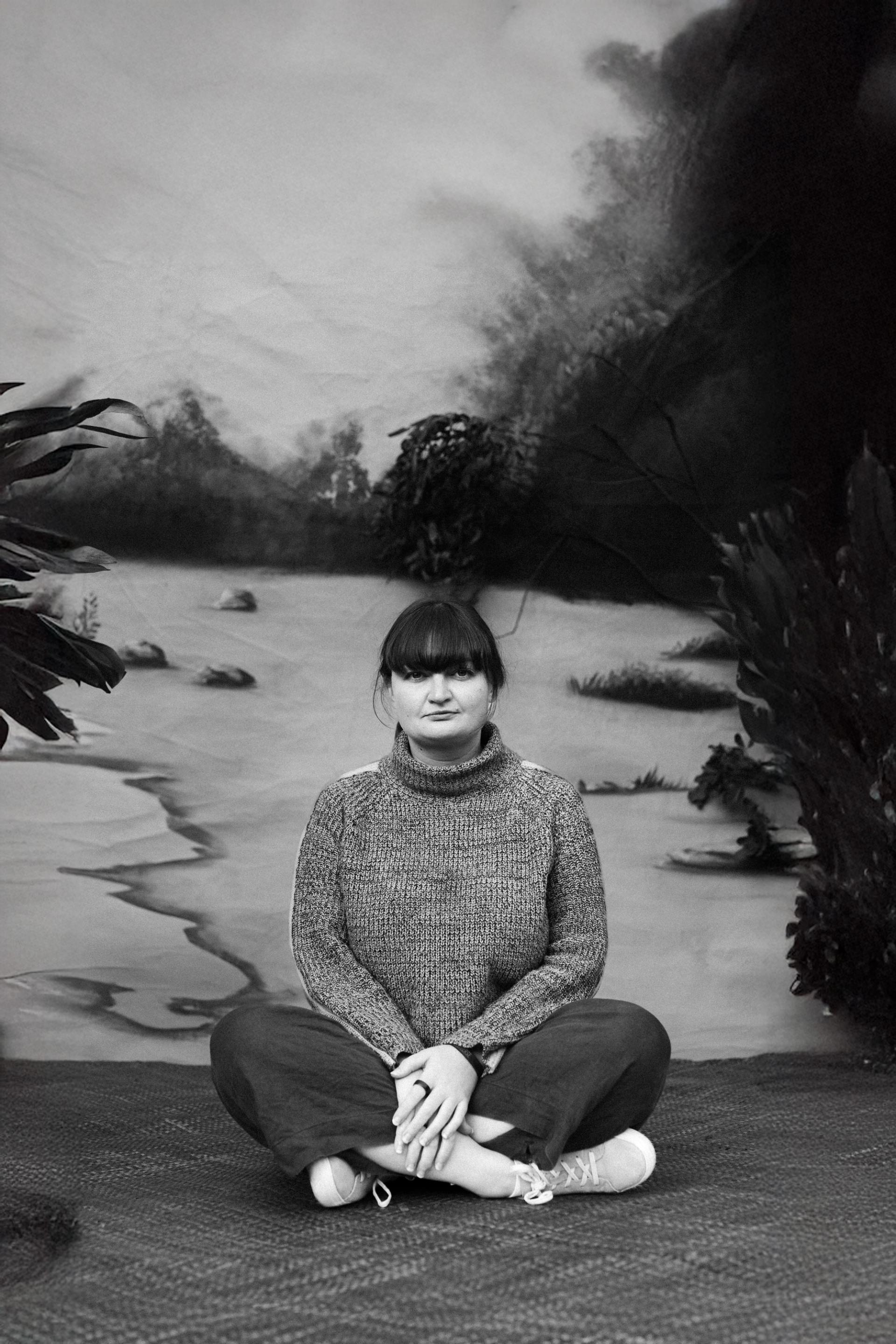 A Palawa woman sits cross-legged on a woven mat. There is a painted backdrop behind her.
