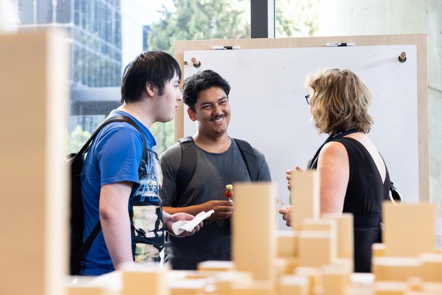 Three humans talking in a circle, in front of a large paper wall, and a wooden model of a city.