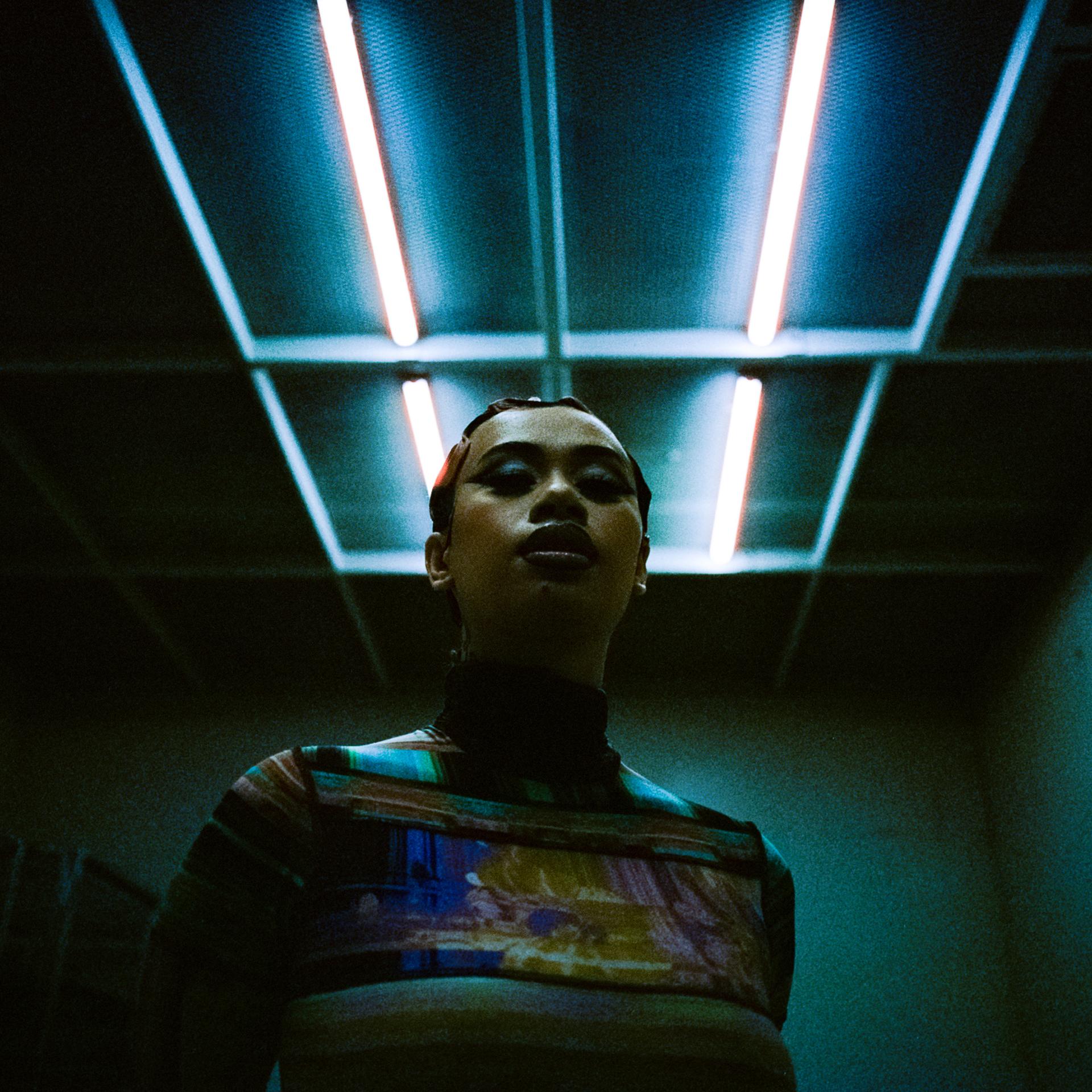 A mid shot of a young woman stands beneath fluorescent light beams.