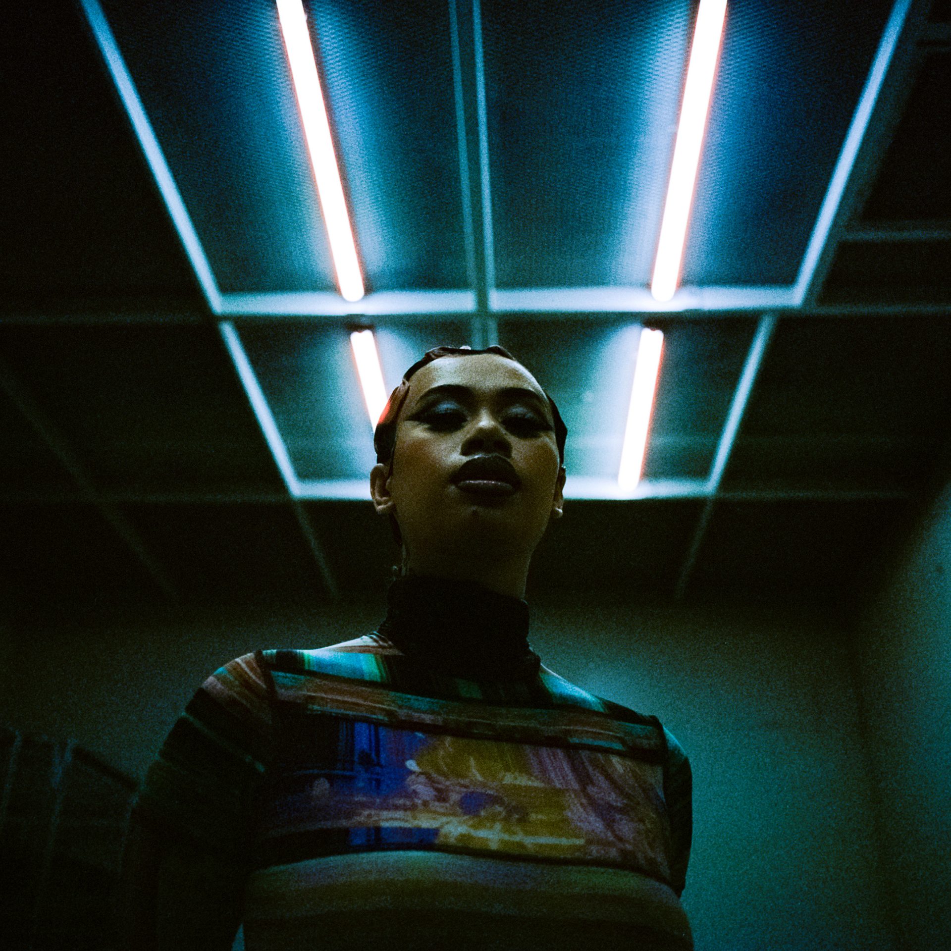 A mid shot of a young woman stands beneath fluorescent light beams.