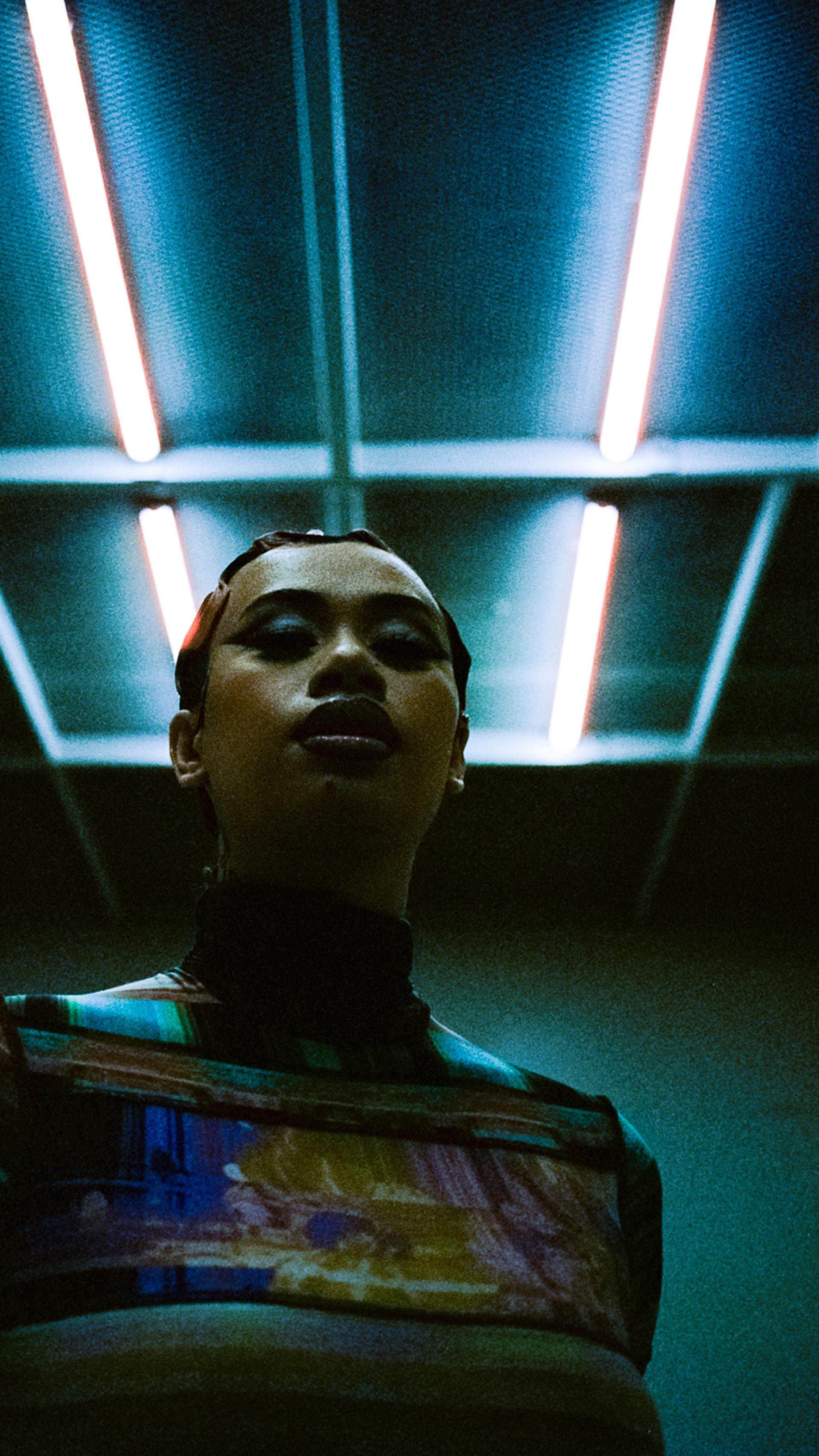 A mid shot of a young woman stands beneath fluorescent light beams.
