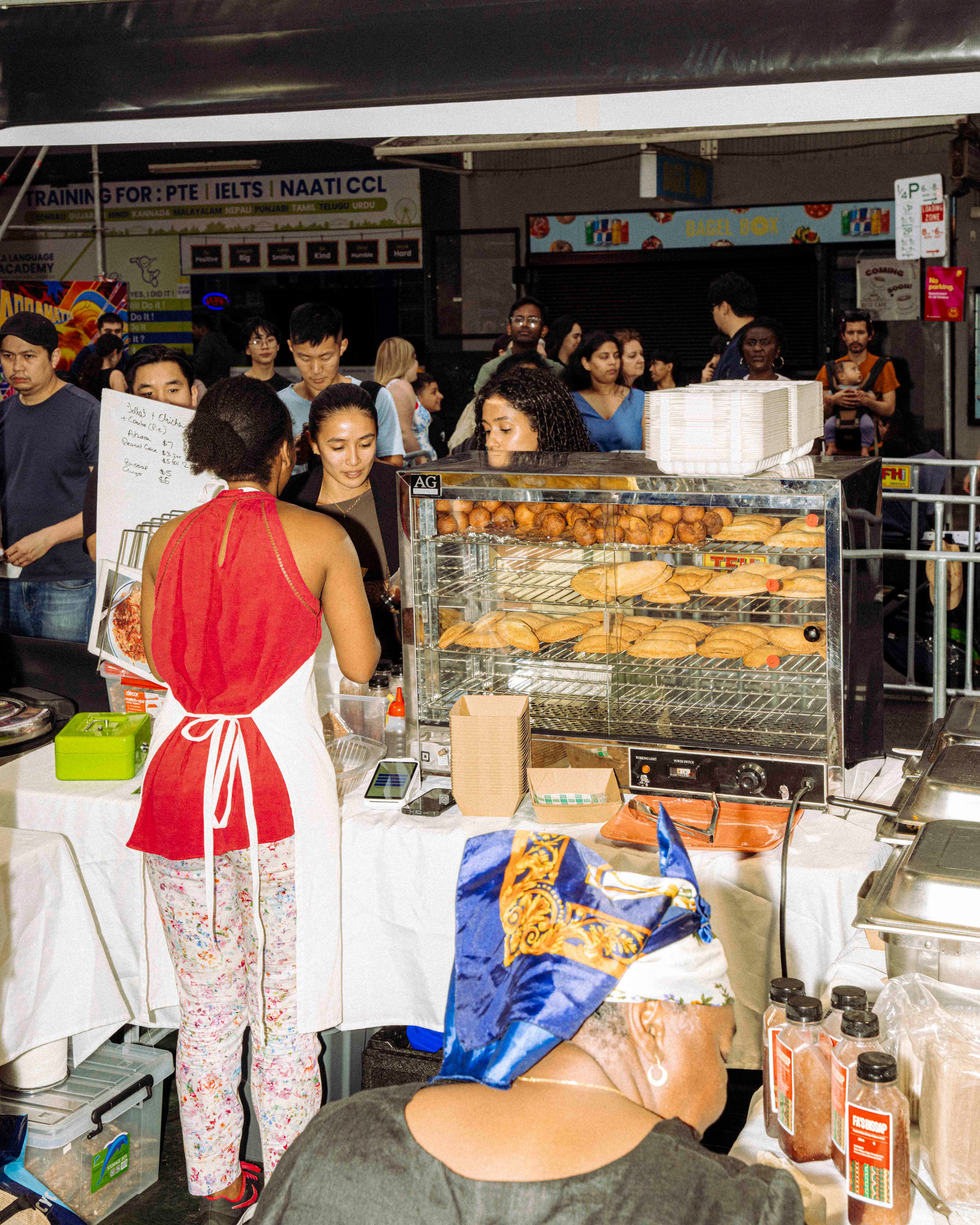 A person in a red top with an apron serves customers at a busy food stall which features a display of pastries. In the background is a diverse crowd of people.