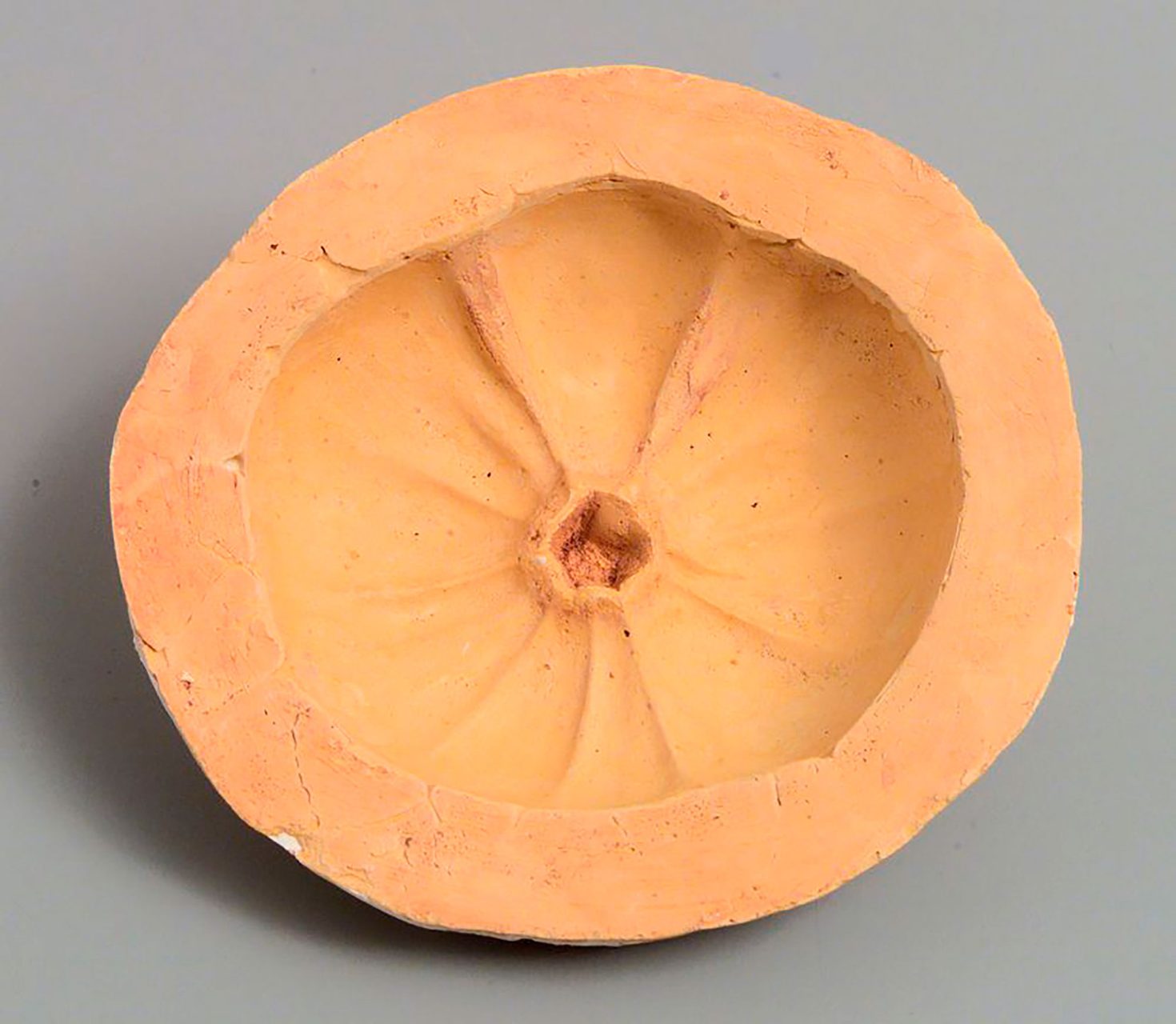 Mould of top half of tomato. Mould side: flattened lip about 20mm wide forms a circumference around hollowed impression of fruit. Hollowed area concave, with a series of ridges running from the edges into the centre of hollow where they intersect with a small raised circular protrusion, pale orange in colour. Outside resembles a meringue, convex with a number of raised swirls, surface uneven and white with orange tinges.