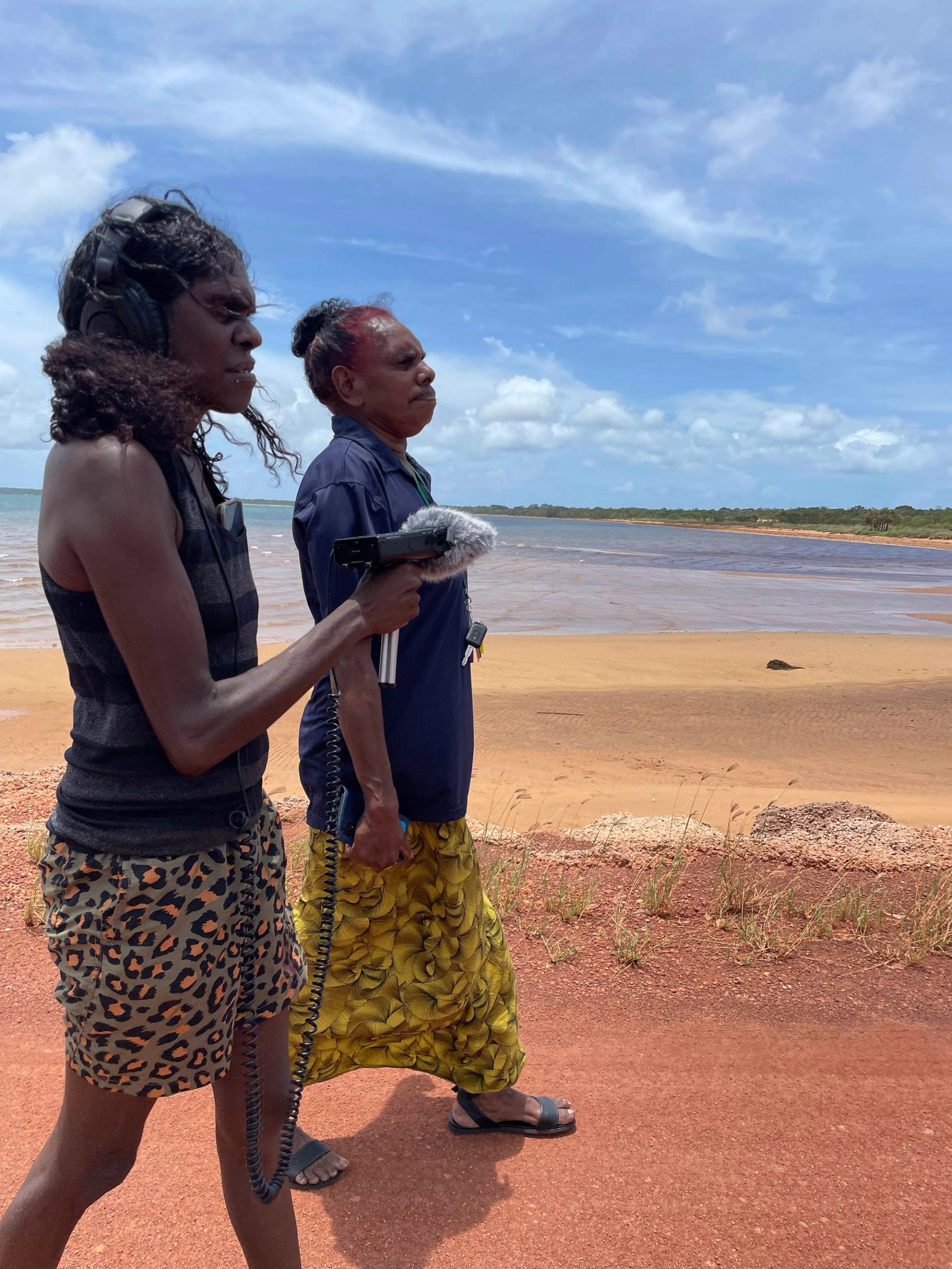 Two women stand in profile, one slightly in front of the other. The woman at the front is holding a microphone and wearing black over ear headphones. They are outside, on the coastline. The earth underneath their feet is red. The ocean is flat, and the sky is blue.