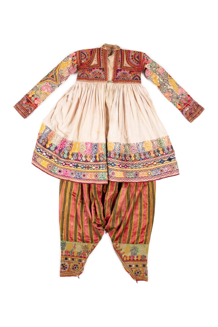 A groom's outfit from the Dhebaria community of Rabari people consisting of headdress (mod) with padded rectangle of beading with silver paper backing, stiffened with 7 sticks, decorated with tufts of multi-coloured wool, a fine red muslin turban (madrasi) with the ends woven in gold metallic, blue, black and white thread in a simple stripe pattern with small geometric motifs, a long sleeved, cream cotton wedding robe is loose fitting with the 'skirt' gathered onto the short yoke, loose fitting trousers (chorano), striped cotton shawl (patolu) in red, black and yellow, heavily beaded sword cover (talvar miyan), and two sets of earrings worn at the top and inner ear (bhungari & toliyo) which are decorative gold metal domes with shanks.