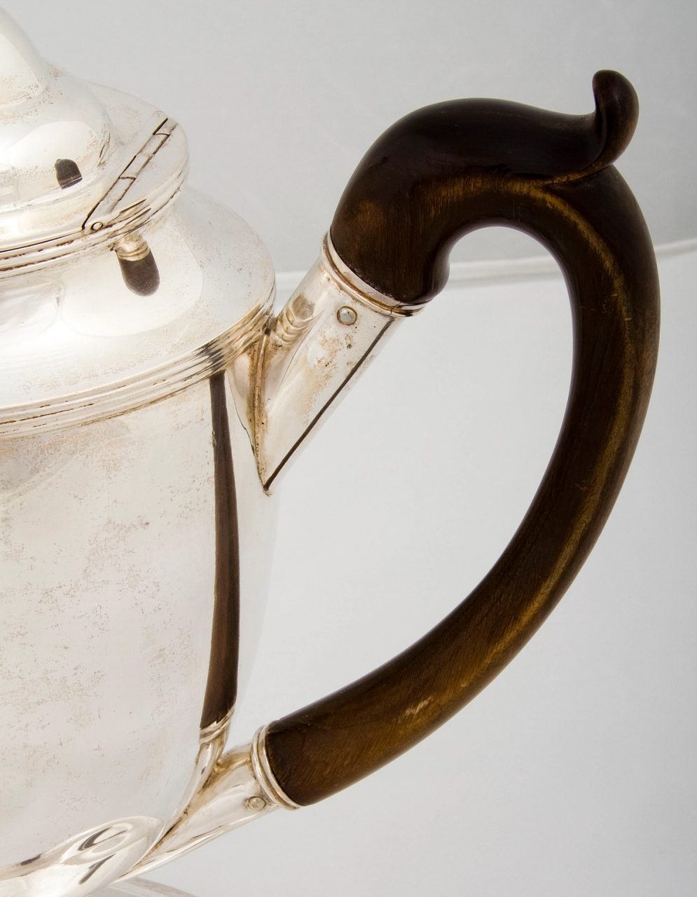 A coffee pot with lid, made of silver and wood, with a c-shaped wooden handle.