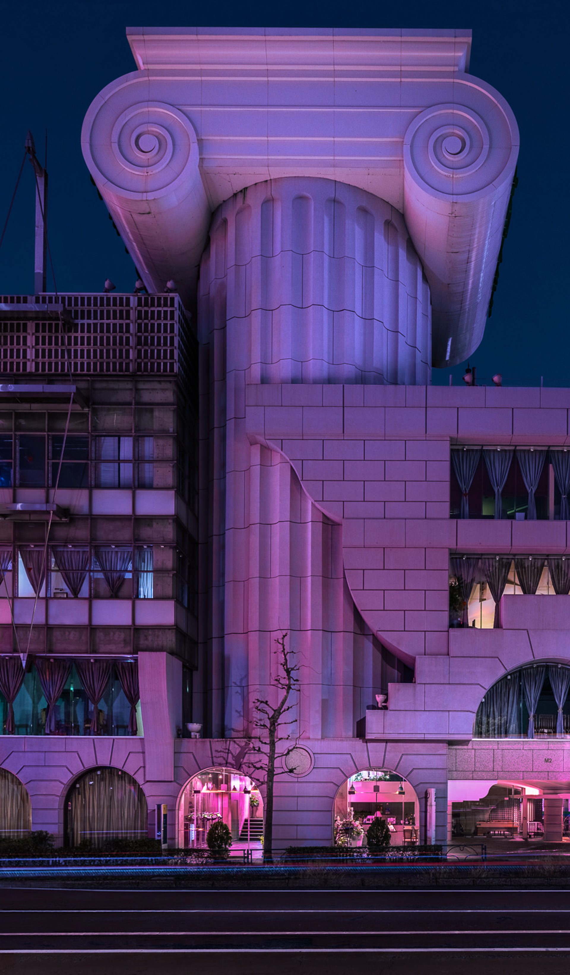 A large, eclectic building at night, lit with purple lights. In the middle of the building is an oversized Ionic column.