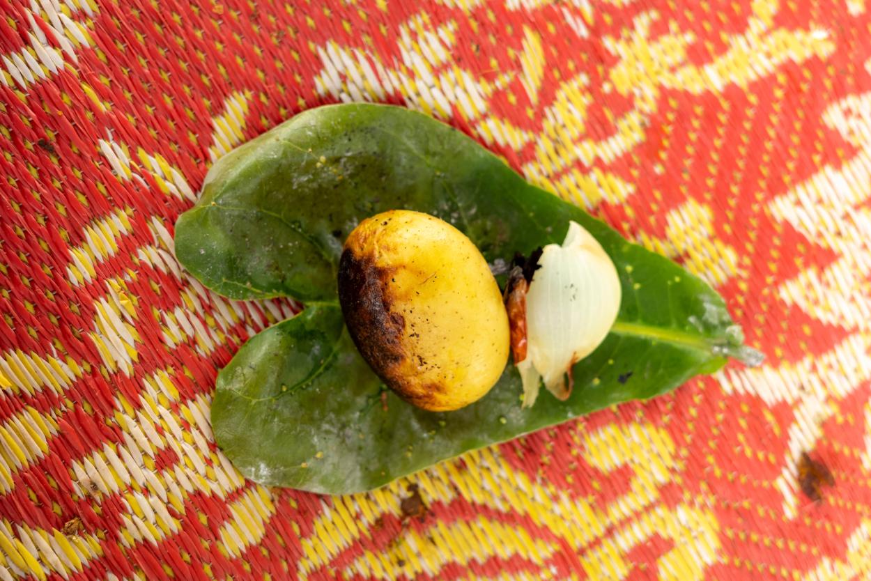 A charred whole potato and piece of cooked onion on a large green leaf, resting on a brightly coloured woven mat.