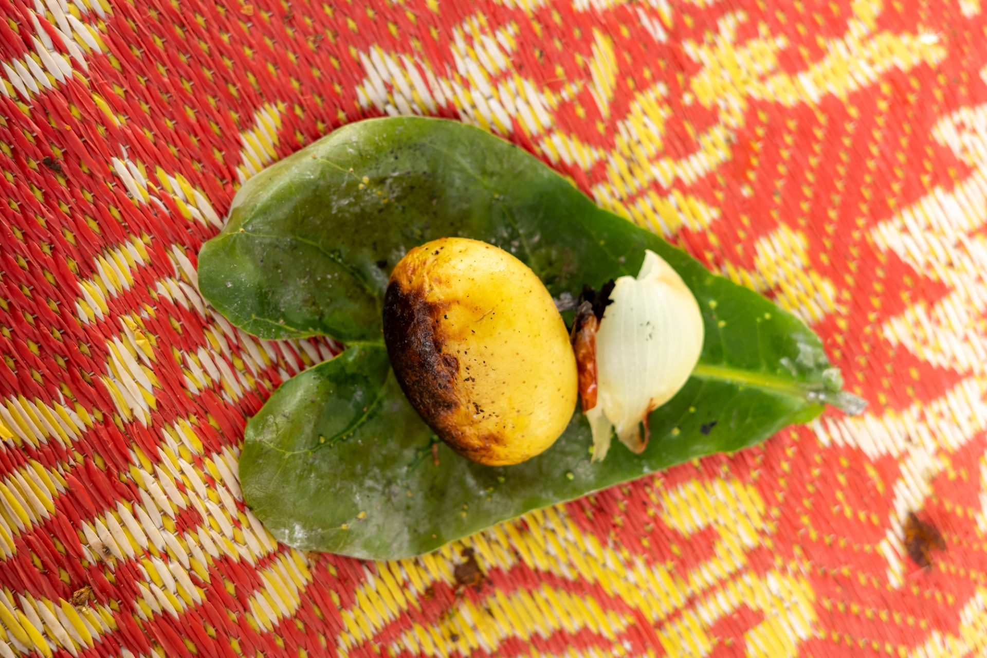 A charred whole potato and piece of cooked onion on a large green leaf, resting on a brightly coloured woven mat.
