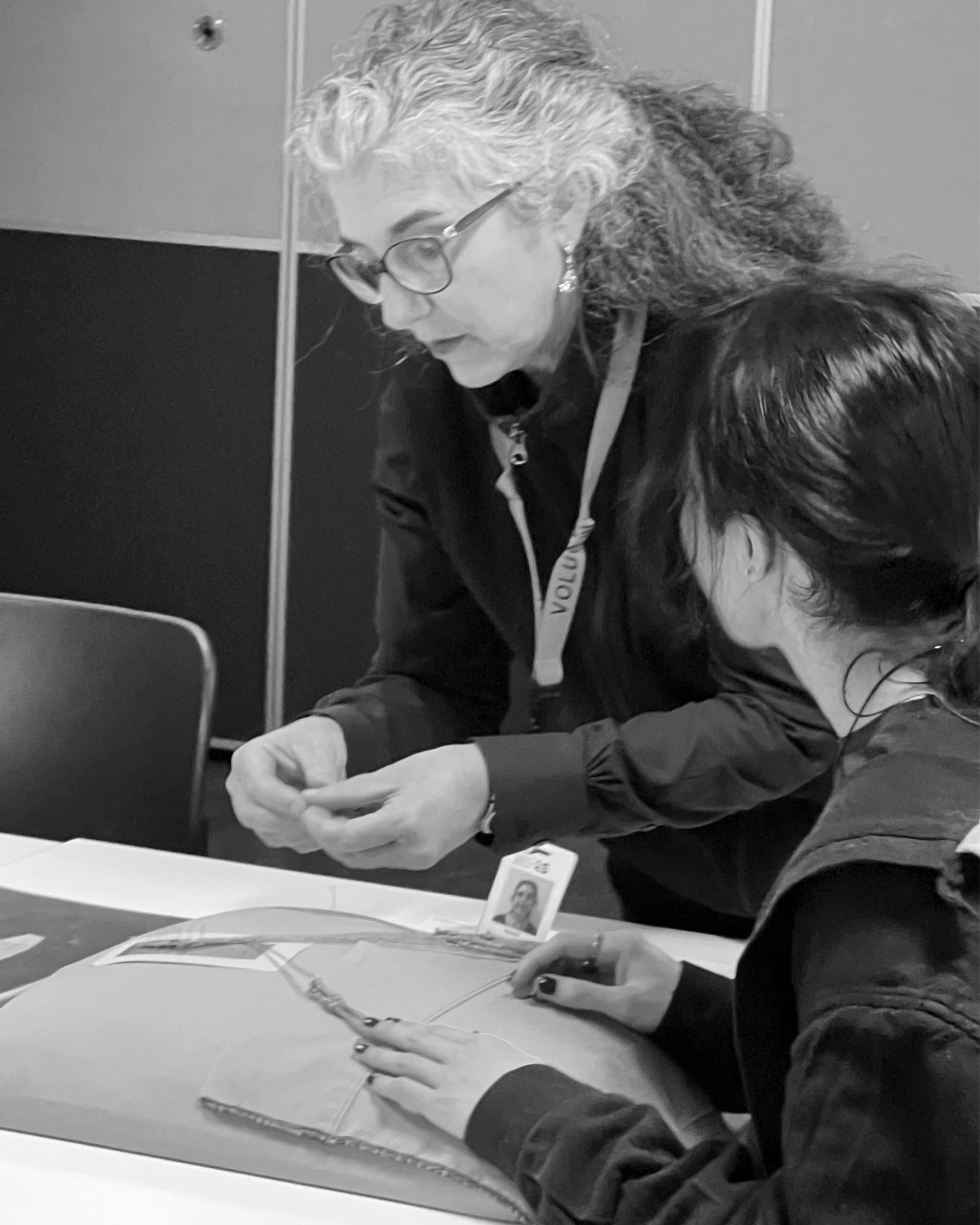 Black and white image of a woman (Isa) showing a student the practice of lacemaking with a lacemaking pillow in front of them.