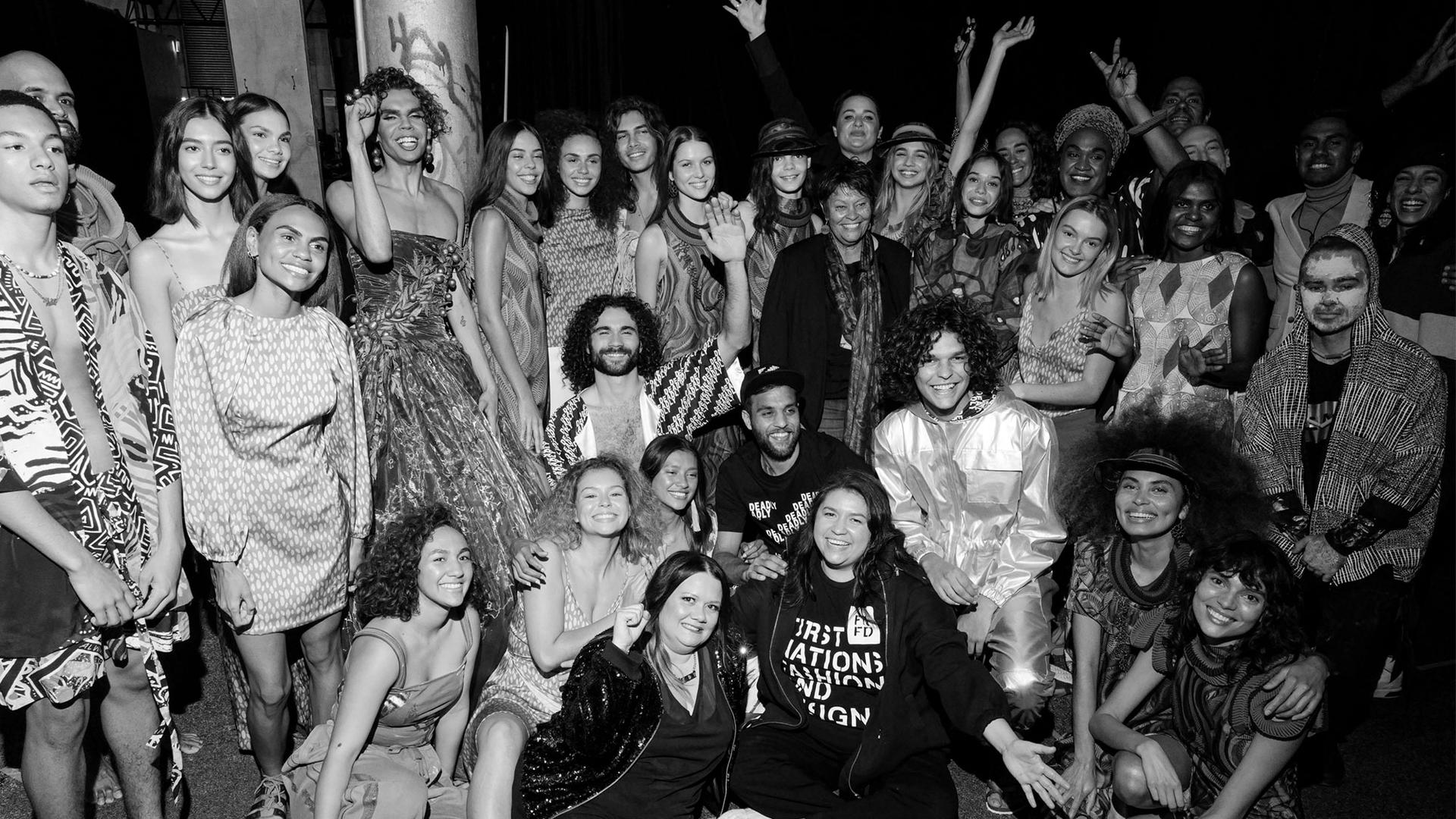 First Nations models dressed in different outfits celebrate backstage at a fashion show. Black and white photograph.