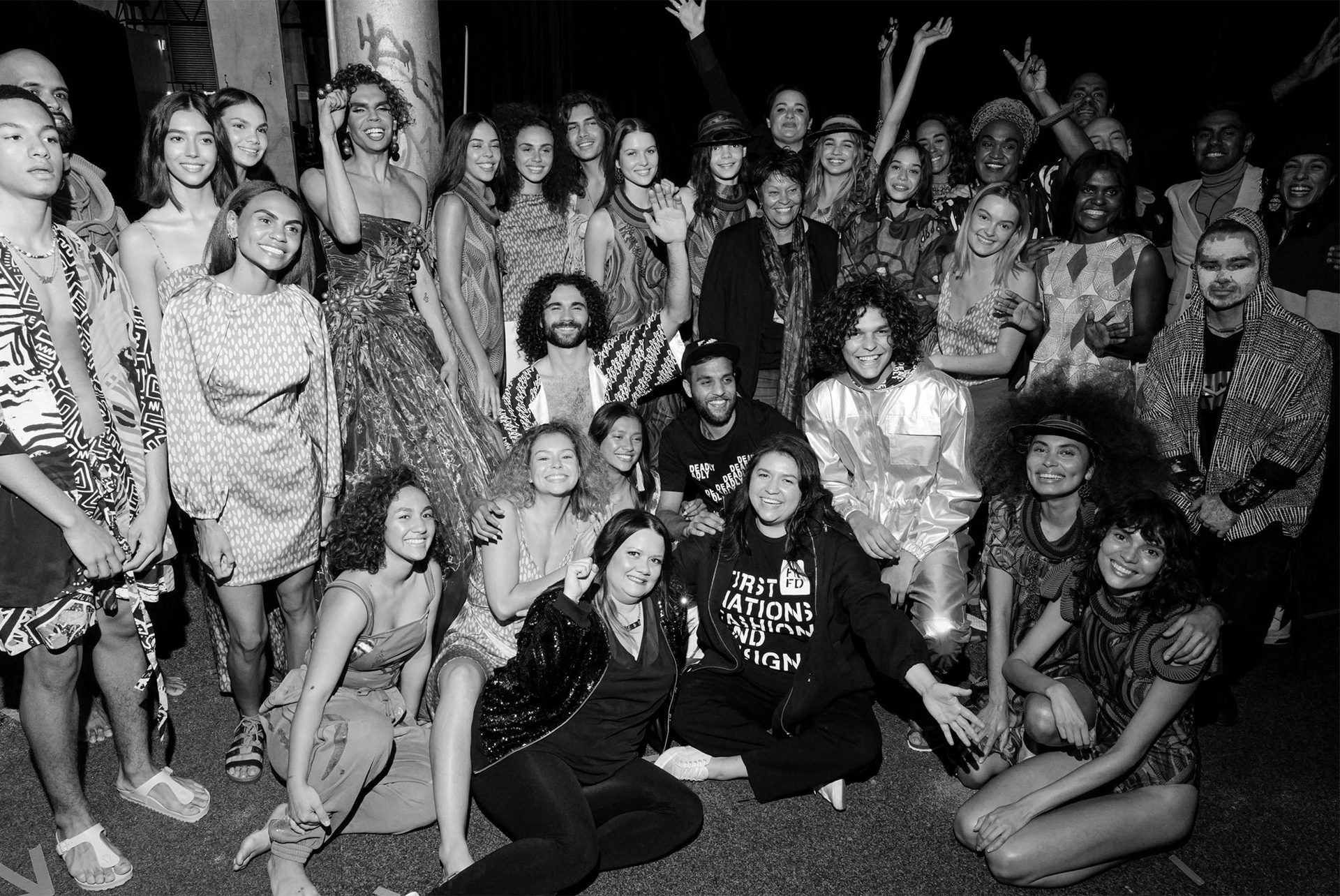 First Nations models dressed in different outfits celebrate backstage at a fashion show. Black and white photograph.