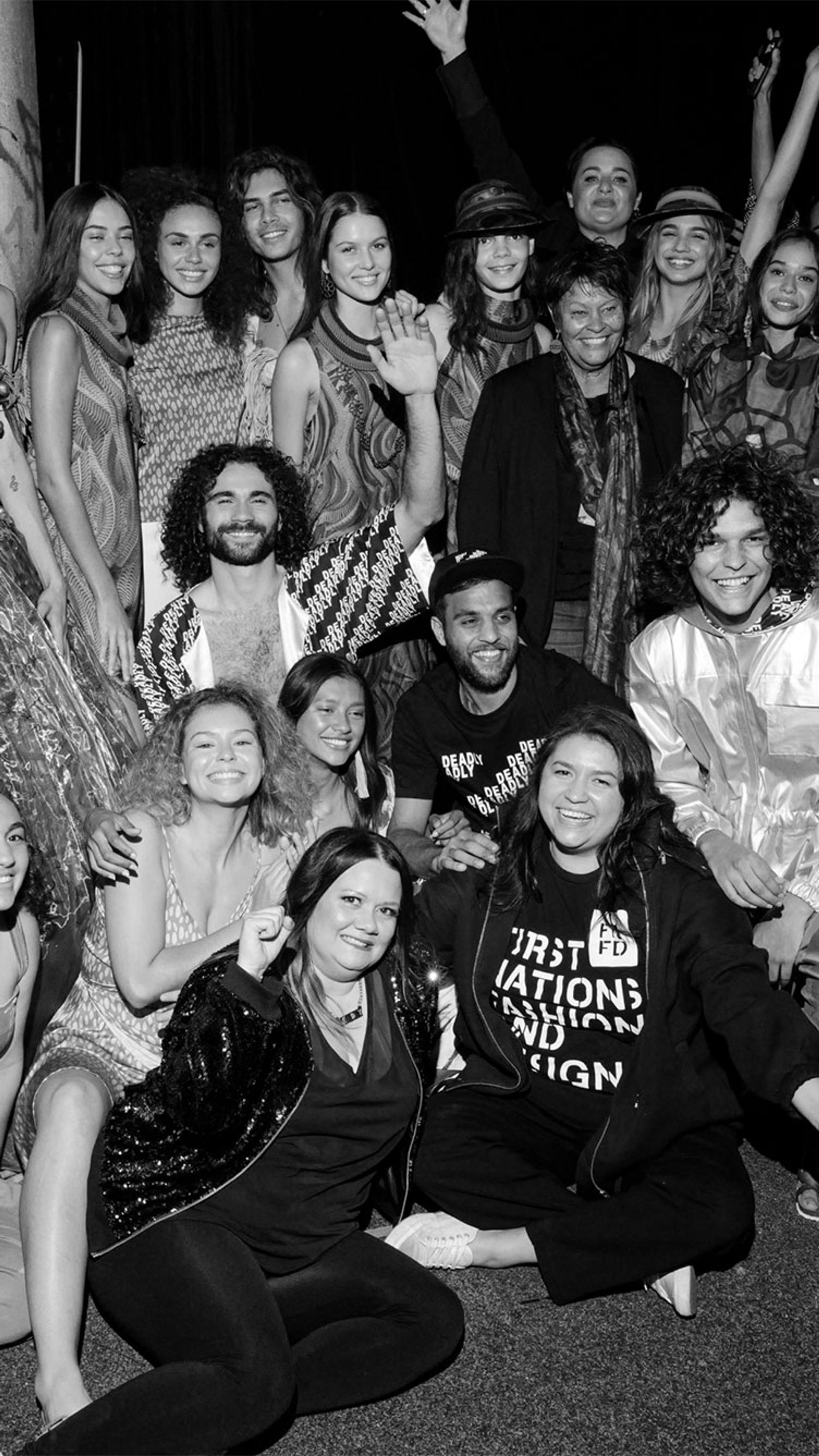 First Nations models dressed in different outfits celebrate backstage at a fashion show. Black and white photograph.