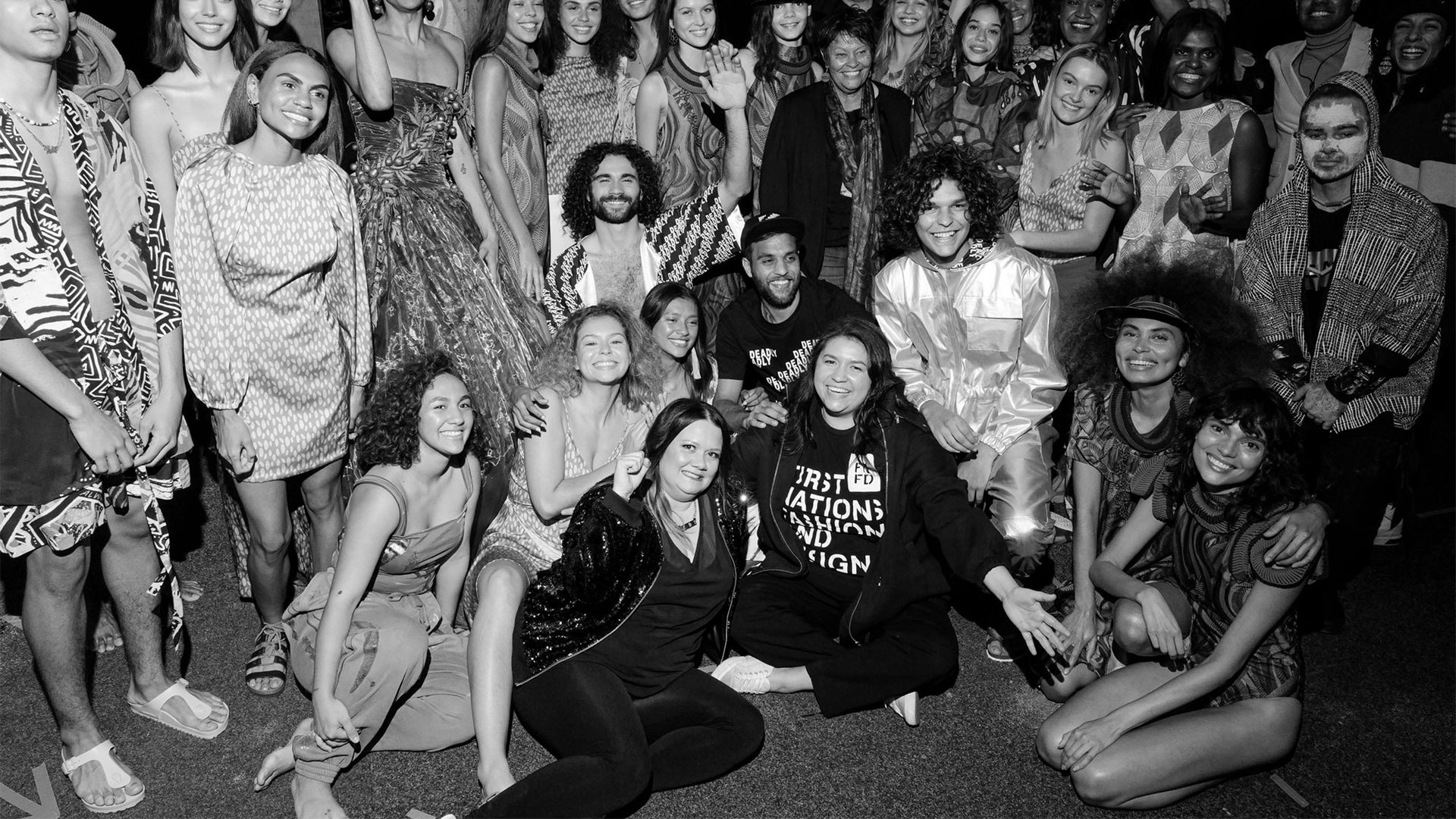 First Nations models dressed in different outfits celebrate backstage at a fashion show. Black and white photograph.