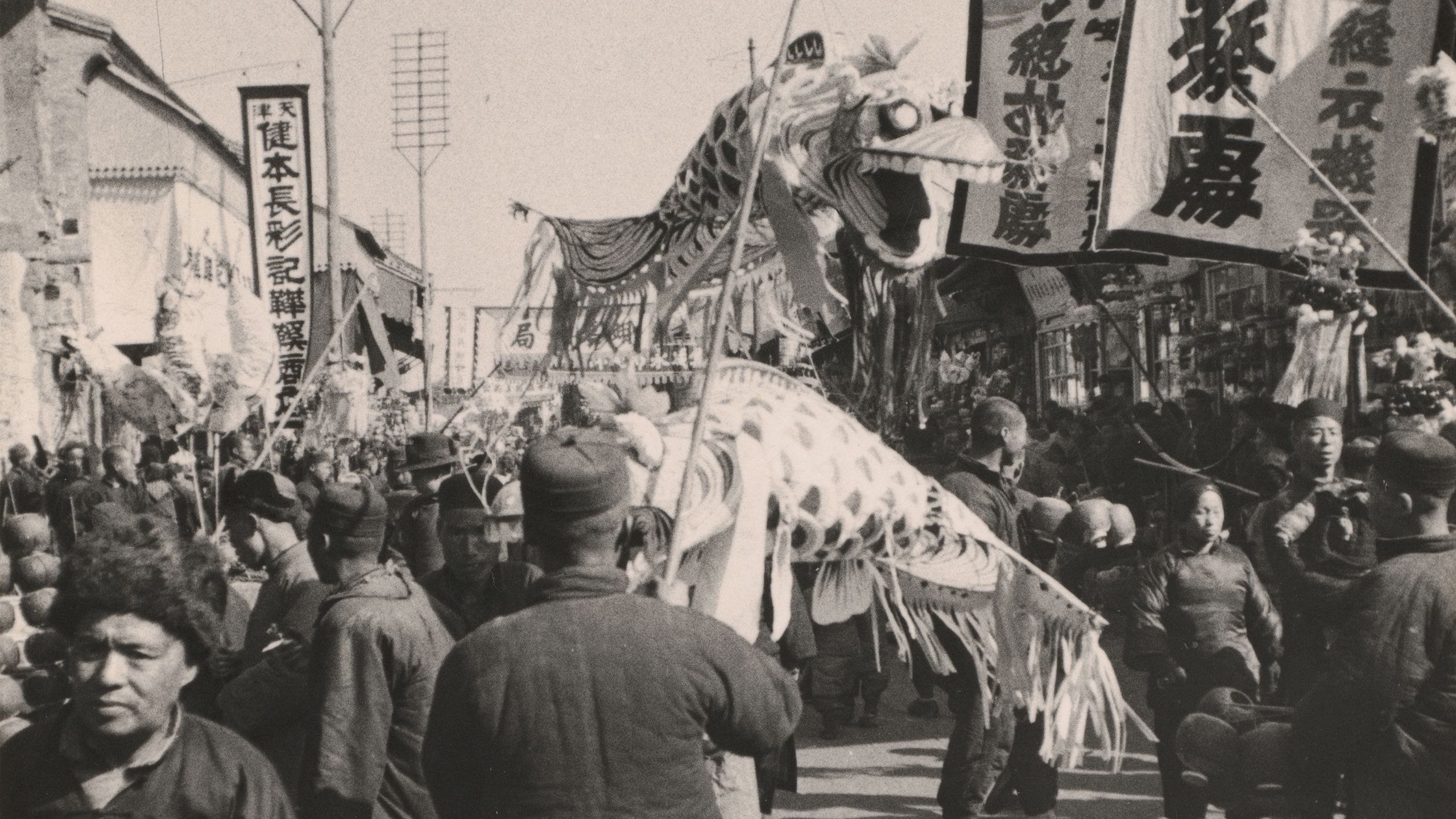Black and white photograph, depicting Chinese New Year Procession. A paper dragon puppet lifted by sticks above a crowd of people, with large banners and signs surrounding them.