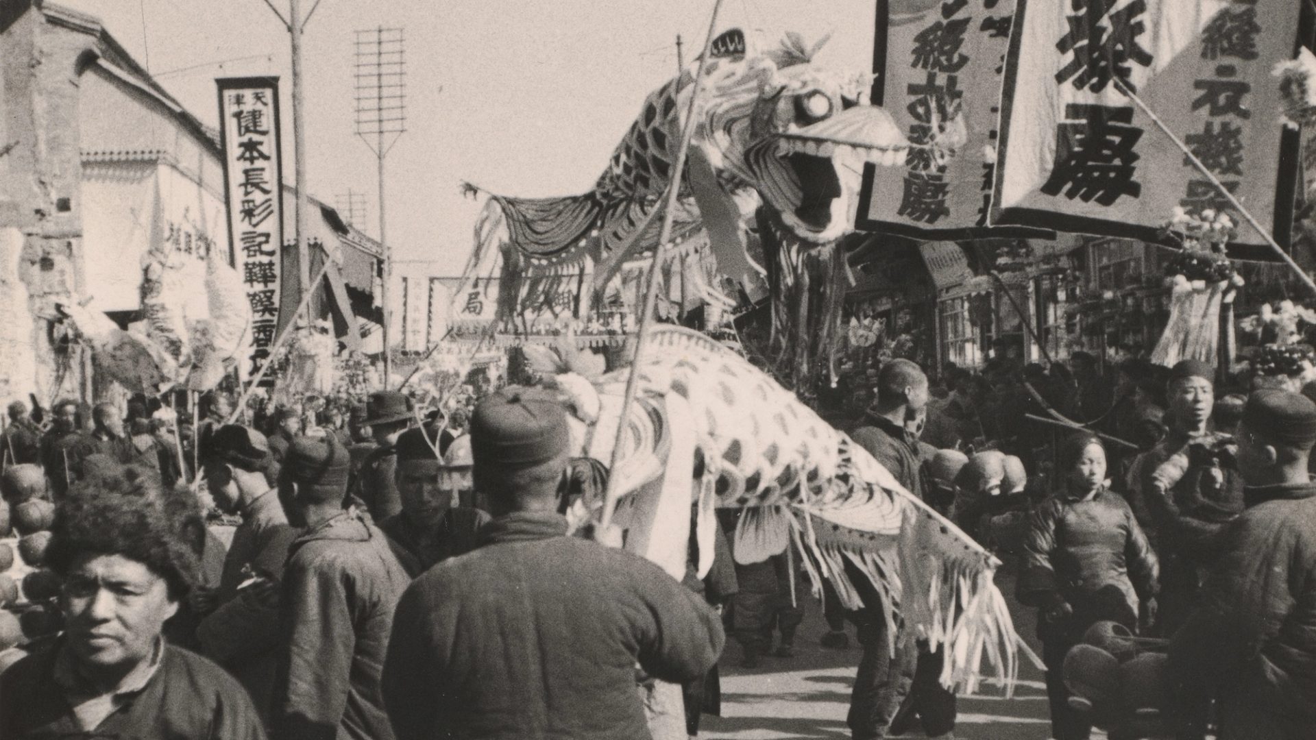 Black and white photograph, depicting Chinese New Year Procession. A paper dragon puppet lifted by sticks above a crowd of people, with large banners and signs surrounding them.