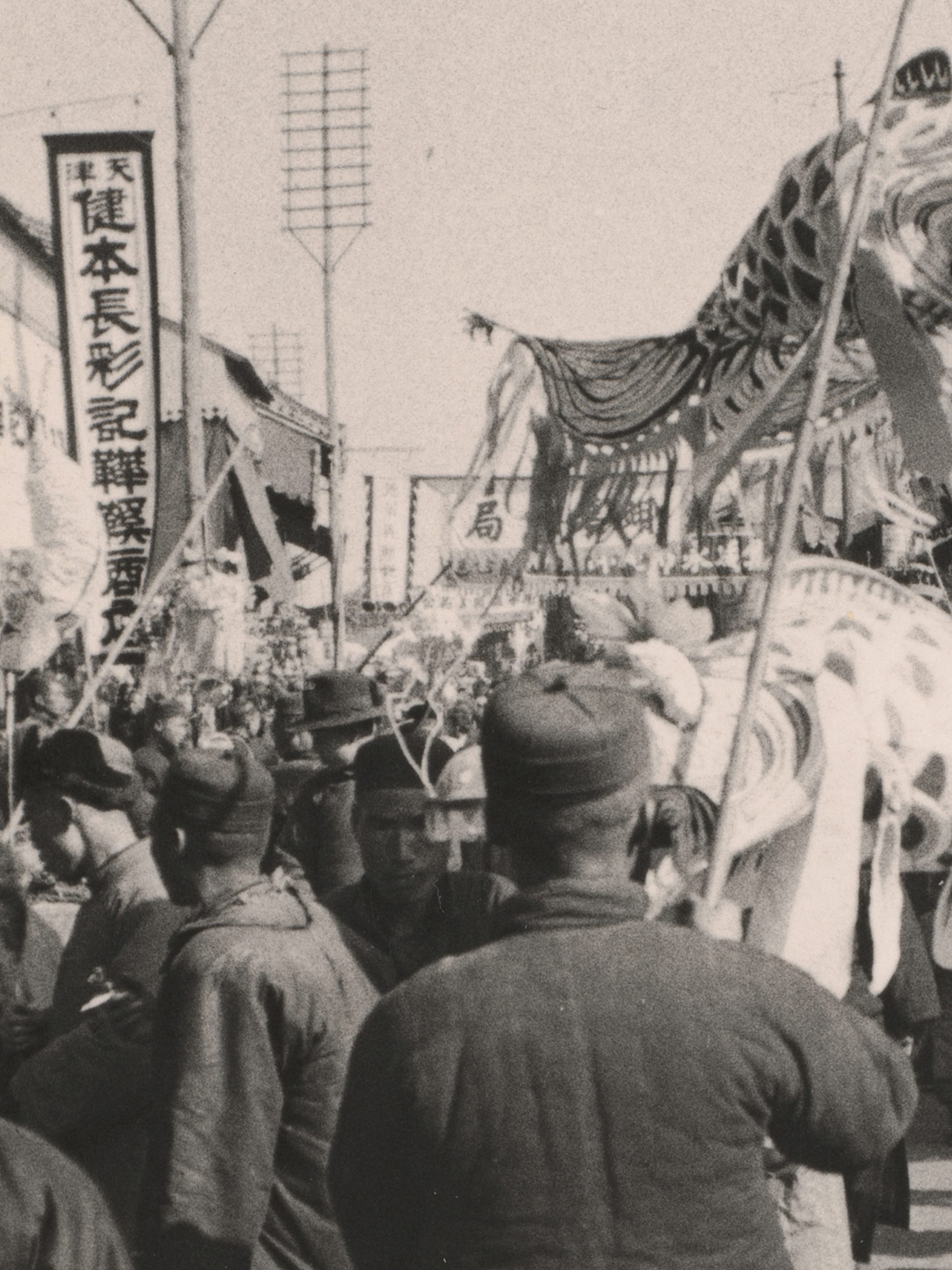 Black and white photograph, depicting Chinese New Year Procession. A paper dragon puppet lifted by sticks above a crowd of people, with large banners and signs surrounding them.