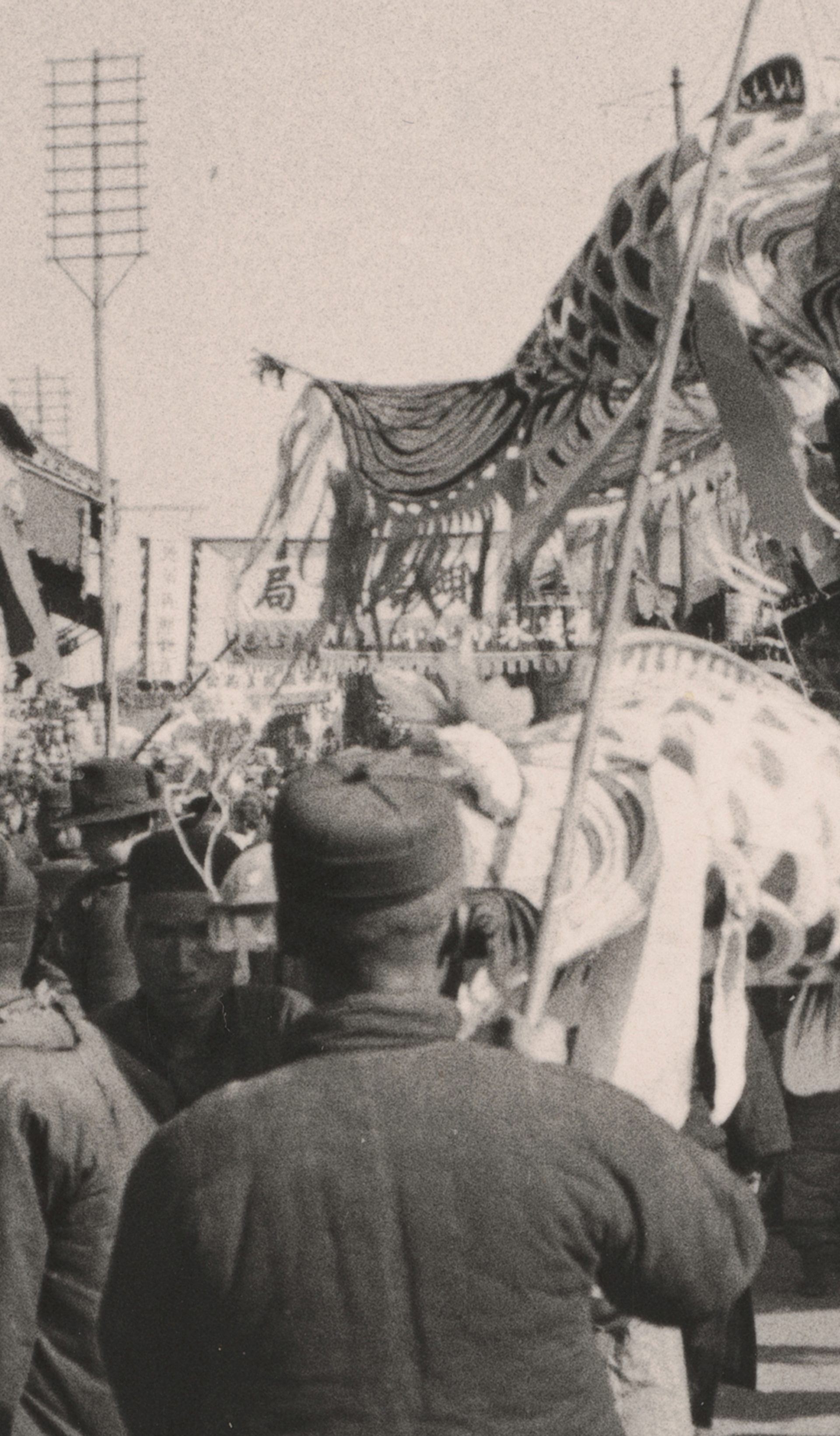 Black and white photograph, depicting Chinese New Year Procession. A paper dragon puppet lifted by sticks above a crowd of people, with large banners and signs surrounding them.