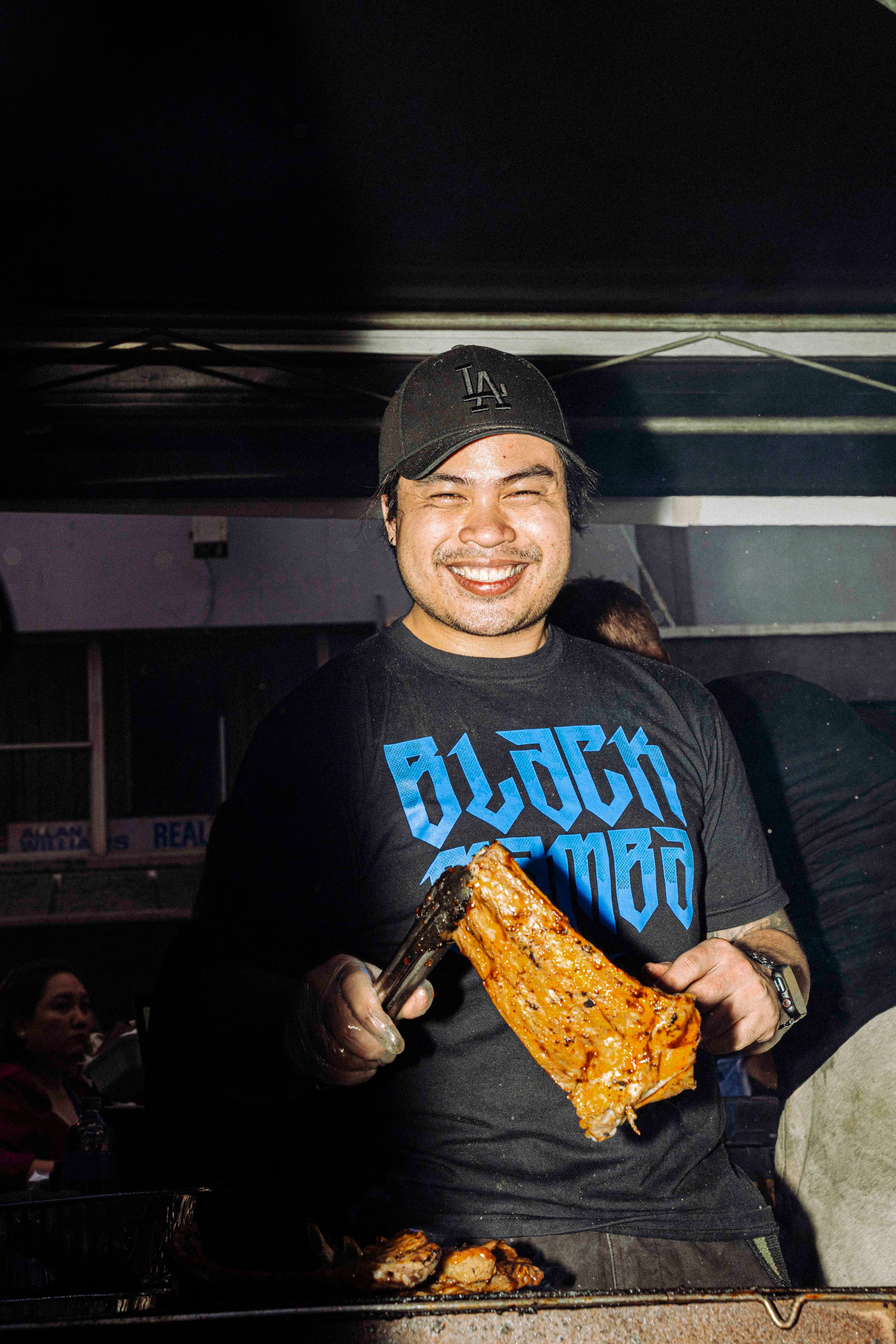 A smiling street vendor wearing a black baseball cap and a blue-lettered ‘BLACK MAMBA’ T-shirt stands, preparing skewers of grilled meat at a food stall.