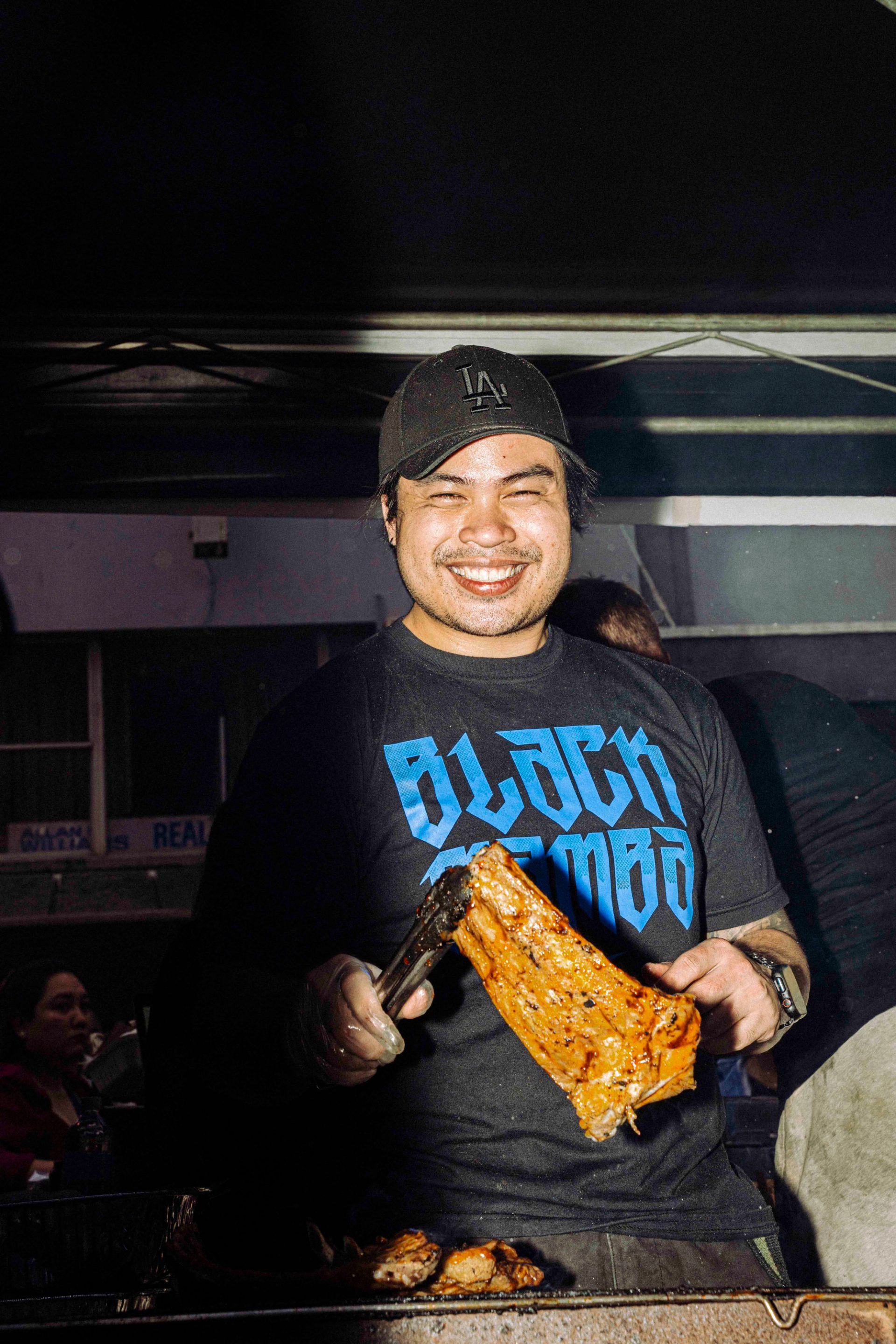 A smiling street vendor wearing a black baseball cap and a blue-lettered ‘BLACK MAMBA’ T-shirt stands, preparing skewers of grilled meat at a food stall.