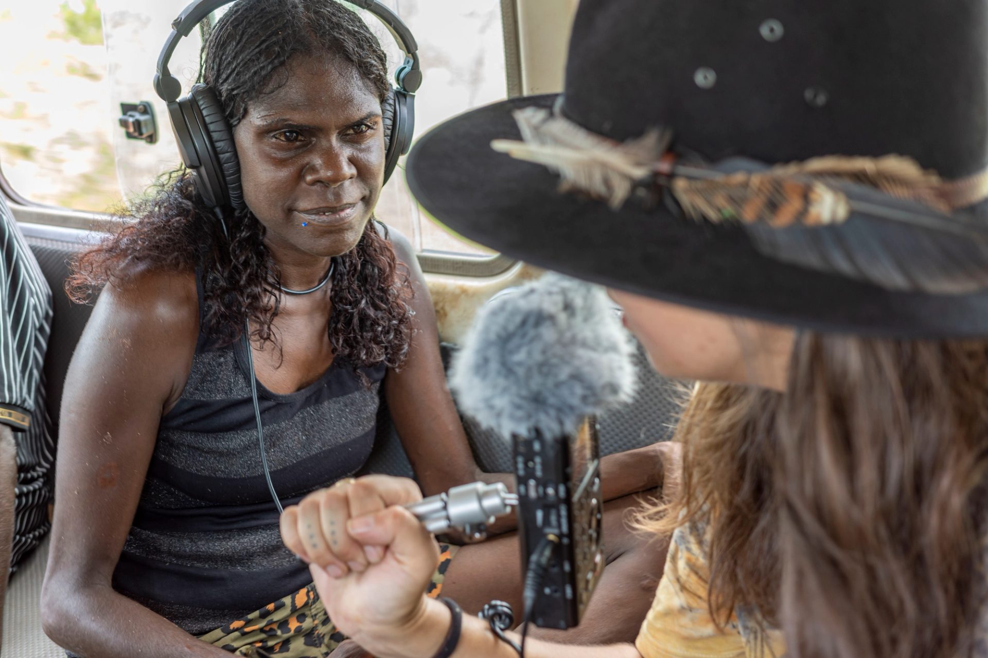 A mid shot of a woman sitting in the back of a troopie wearing a pair of black over ear headphones and a black and grey striped tank top. In the foreground is the side profile of the interviewer. She is wearing a black hat with a feather band and is holding a microphone.