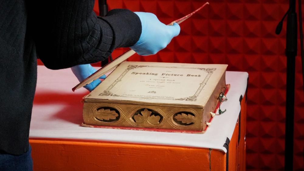 Hands wearing blue plastic gloves hold the cover of a large book-shaped music box. The music box sits on an orange box with a white padded top inside an orange sound booth.