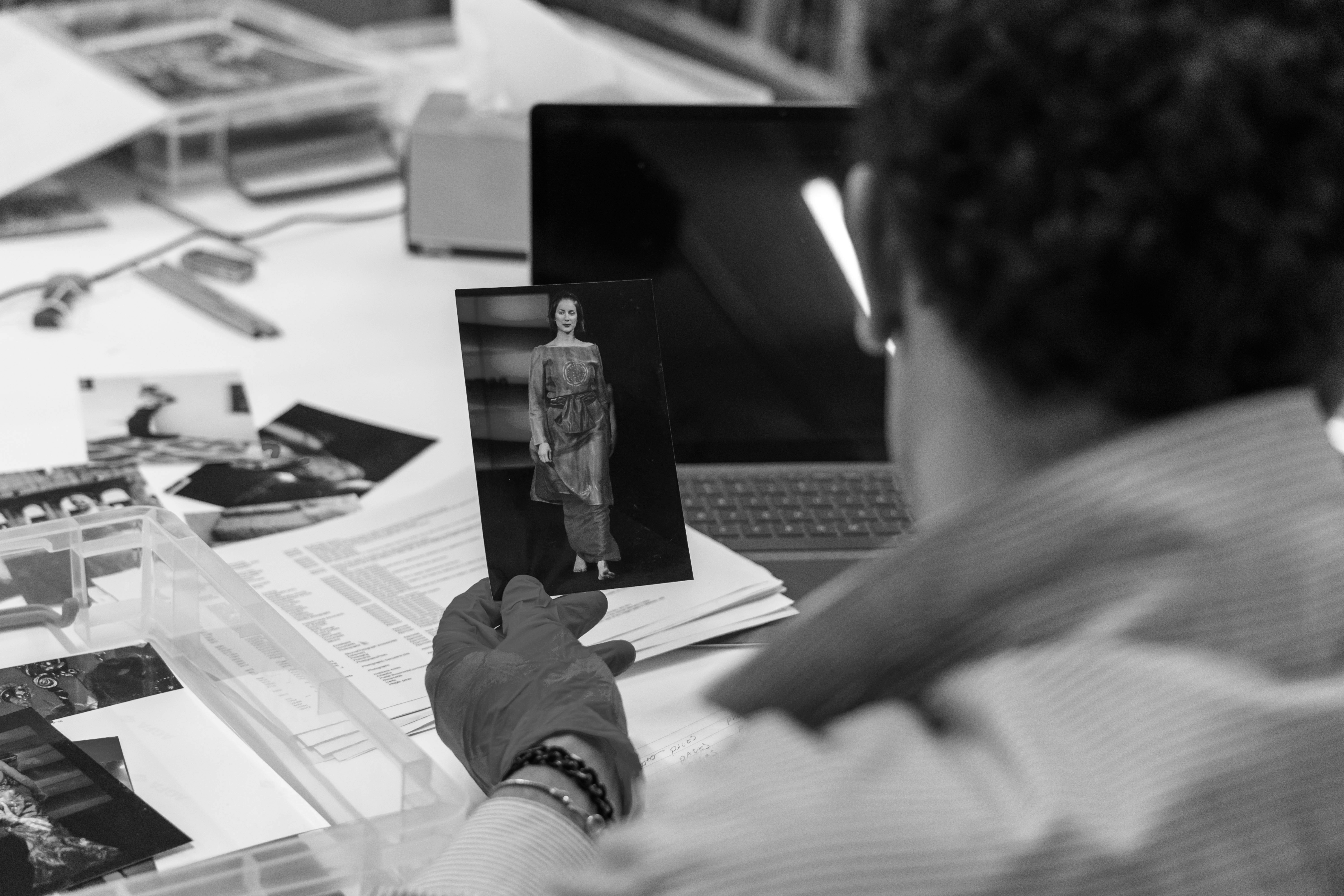 Black and white image looking over the shoulder of a person with curly hair, holding a printed image of a fashion runway model. Background shows more photographs, documents and a laptop scattered across the table in front of the person.