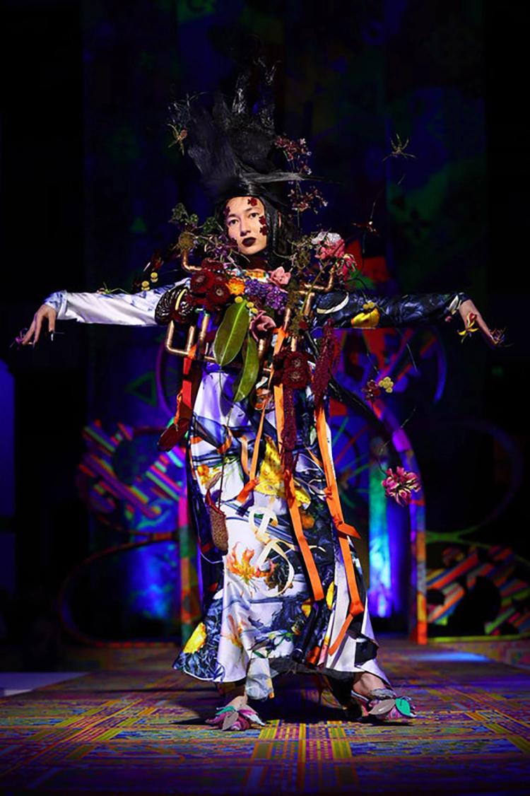 A model poses on the runway wearing a floor-length multicoloured dress that features a flower pattern, and a elaborate sculptural vest made of copper pipes holding an array of flowers and leaves.
