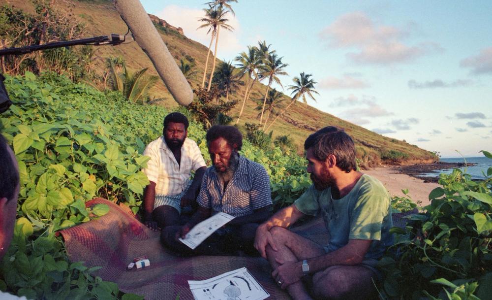 Three men sit on a rug in vegetation just above a beach. They are looking at documents. There is a pack of cigarettes and a lighter on the rug and an audio boom above.