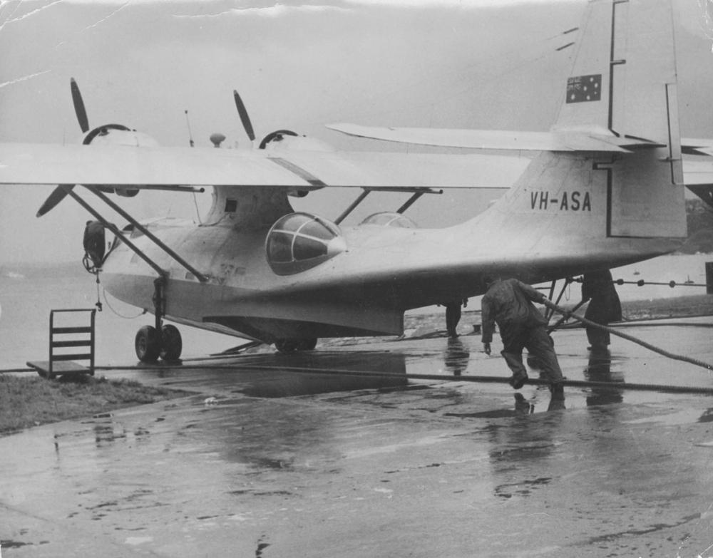 Sliver plane sits on a boat ramp. Two men are attaching ropes to it. Black and white photograph.