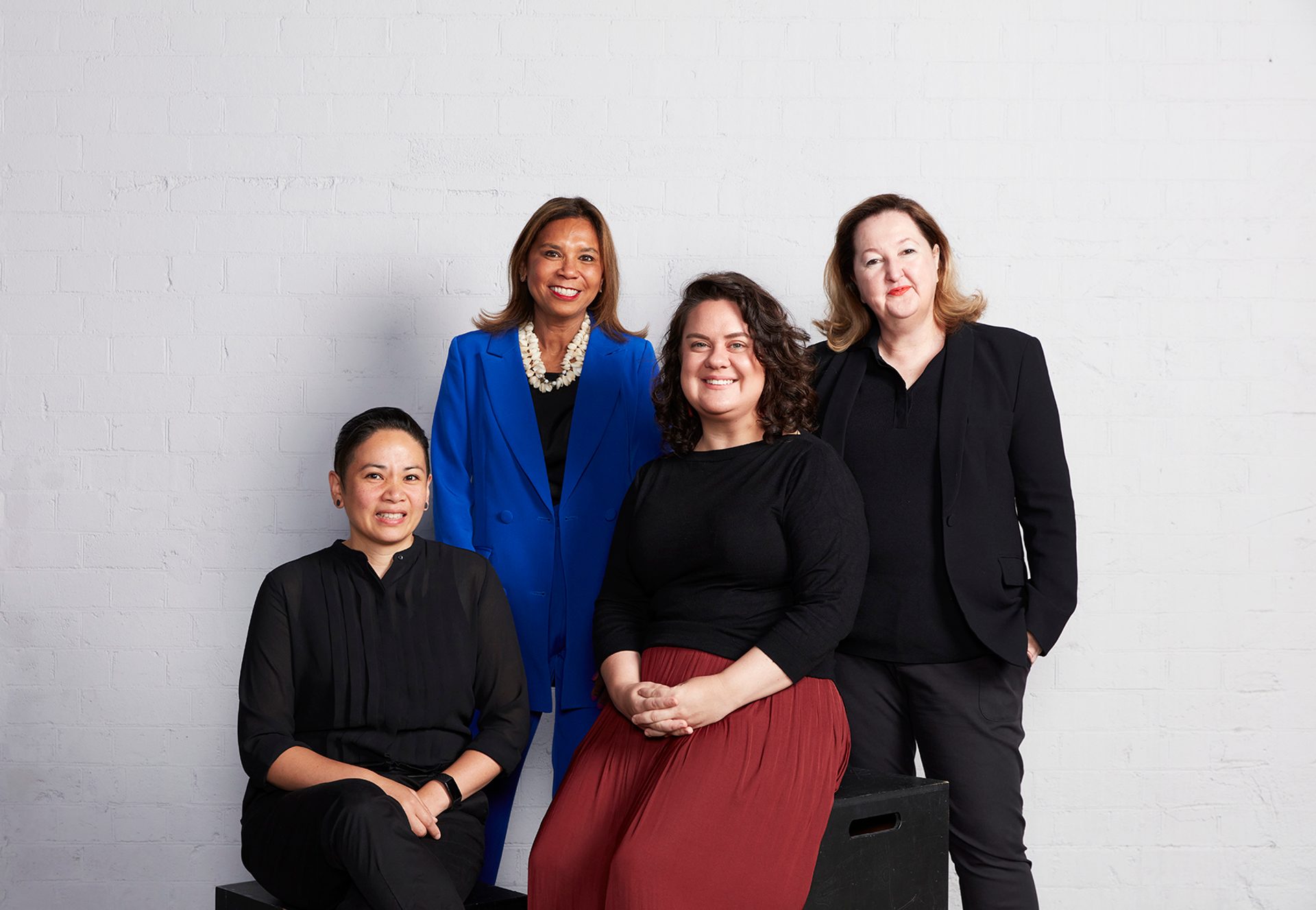 Four figures wearing black, blue and red standing together for a photograph against a white brick wall background.