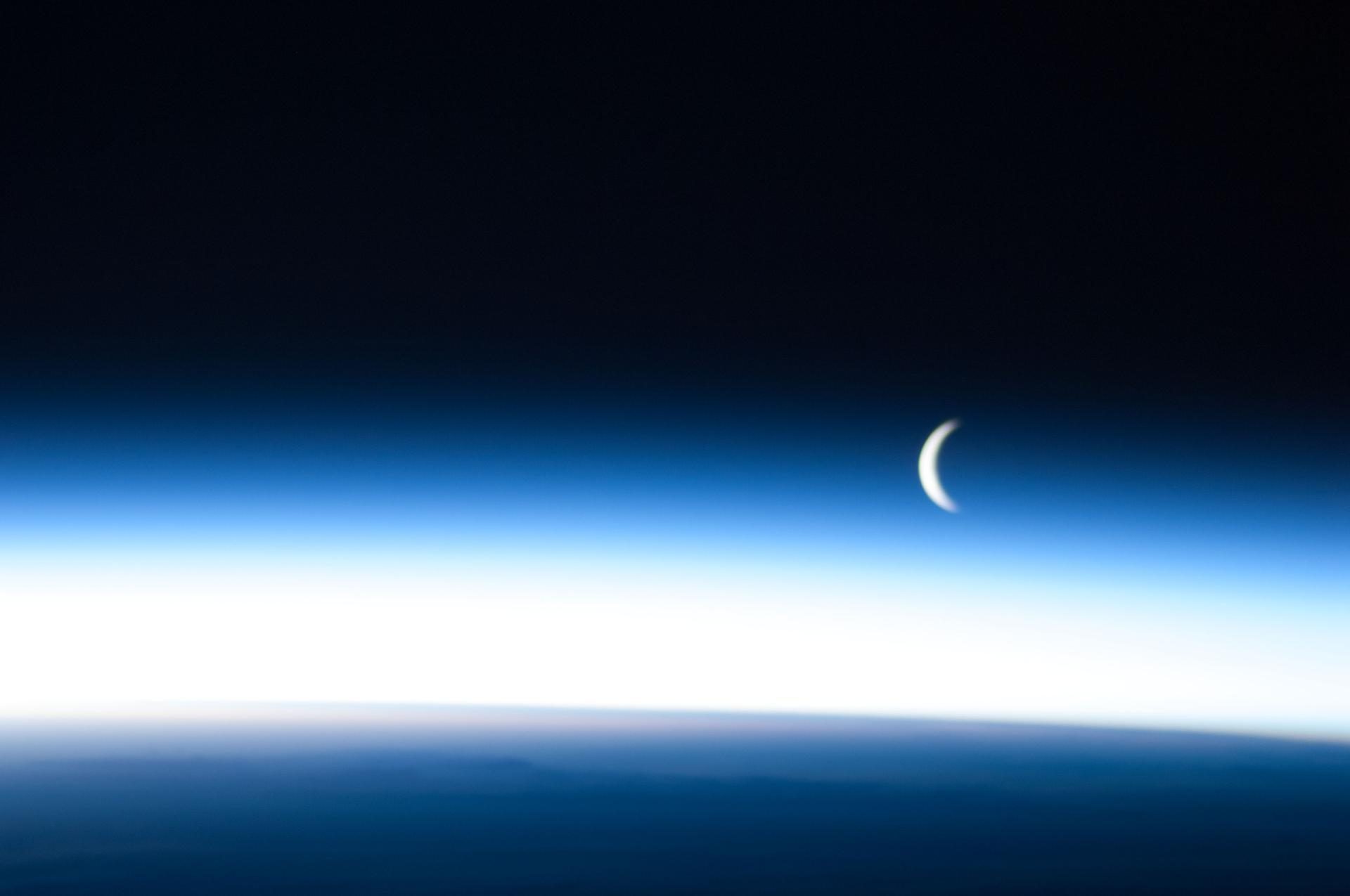 Crescent moon above Earth's atmosphere lit with blue.