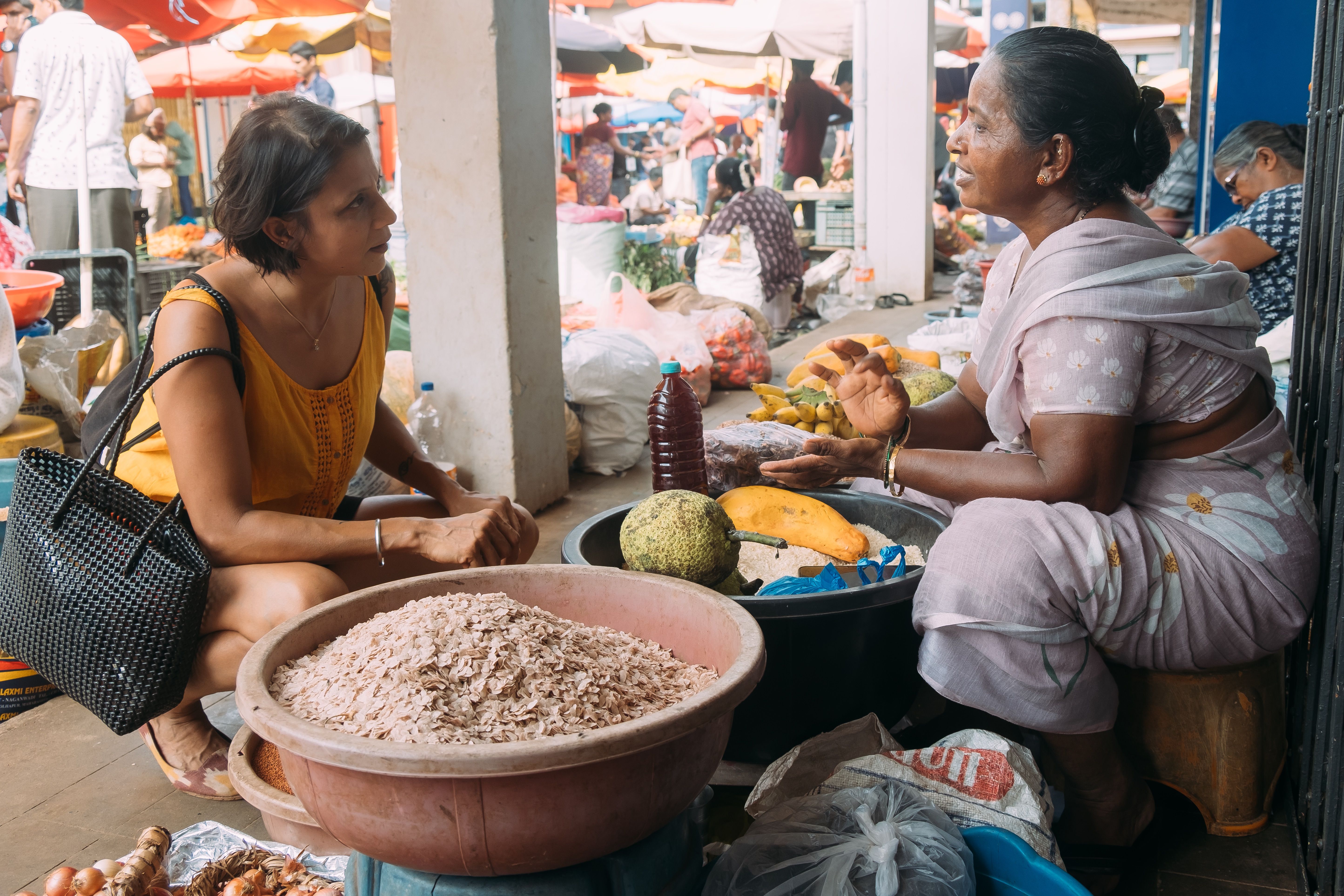 In a local Indian fresh produce market, a female chef crouches while in conversation with a female stall holder.