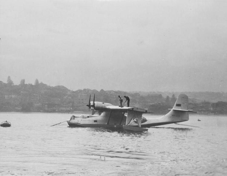 Silver Catalina flying boat is on the water. Two men are standing on the wing cleaning it. Black and white photograph.